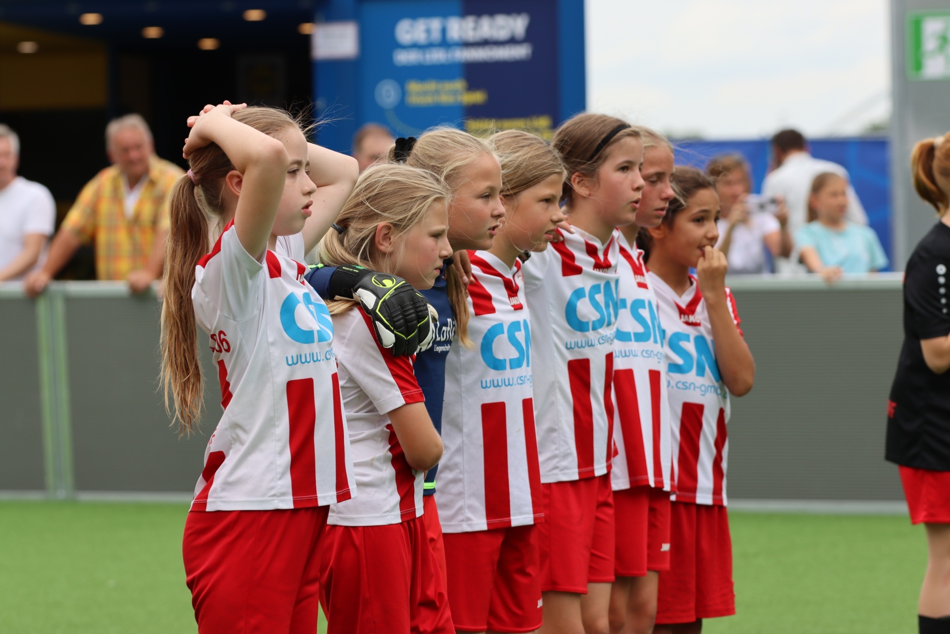 [{"clang_id":1,"value":"Fußballmannschaft aus jungen Mädchen in roten und weißen Trikots steht in einer Reihe und hält sich die Stirn. Im Hintergrund Zuschauer und ein Banner."}]