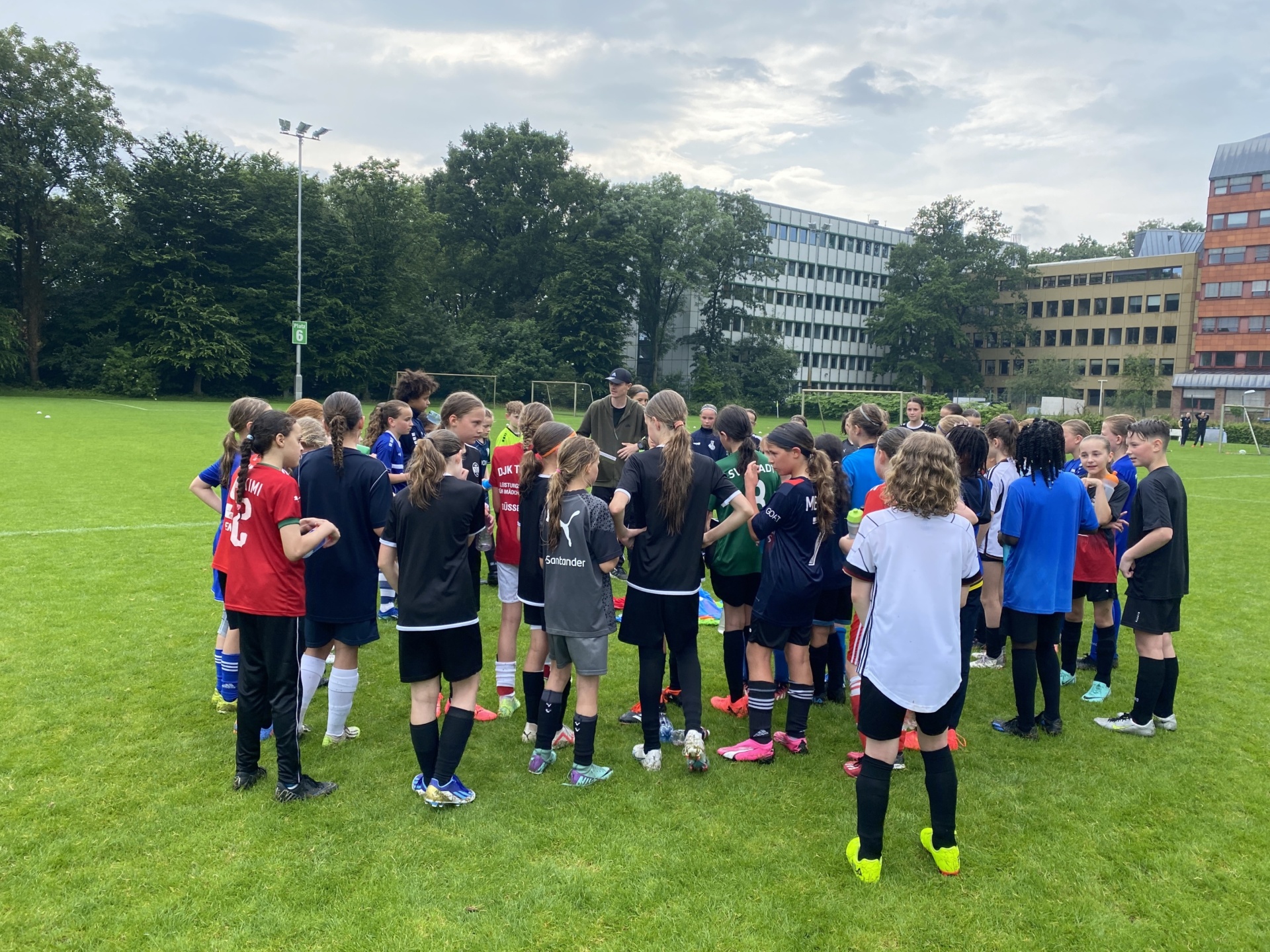 [{"clang_id":1,"value":"Fußballspielerinnen und -spieler in verschiedenen Trikots stehen in einem Kreis auf einem grünen Rasenfeld. Im Hintergrund ein Gebäude und Bäume unter einem bewölkten Himmel."}]