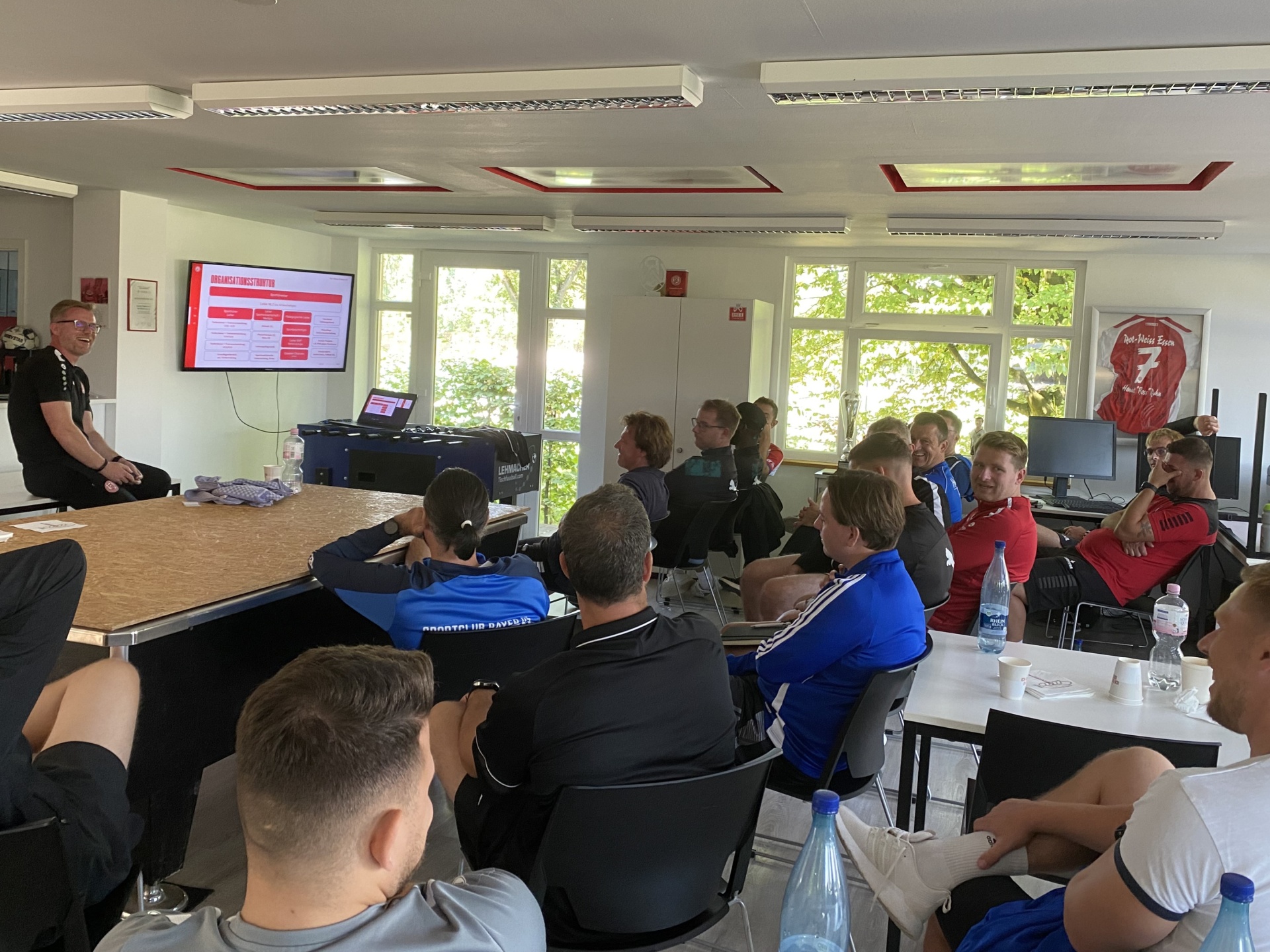 [{"clang_id":1,"value":"Raum mit Präsentation. Gruppe von Männern sitzt an Tischen, ein Mann steht vorne und spricht. Fenster mit Blick auf Bäume im Hintergrund."}]