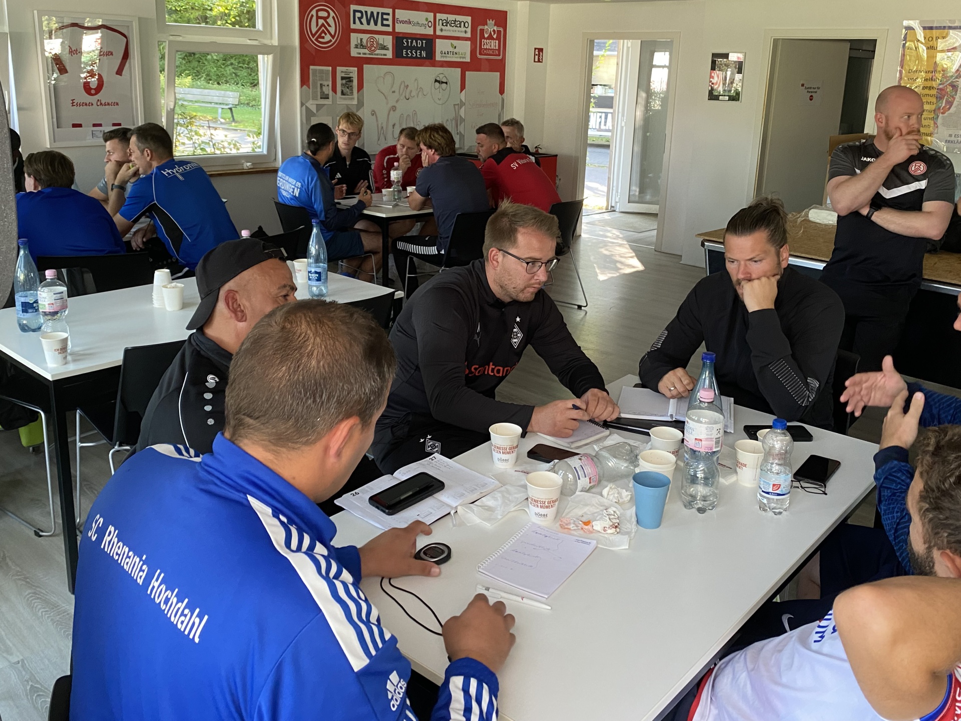 [{"clang_id":1,"value":"Gruppe Männer sitzt an Tischen in einem Raum, vermutlich ein Vereinsheim. Einige schauen auf Smartphones, andere unterhalten sich. Im Hintergrund Vereinsbanner."}]