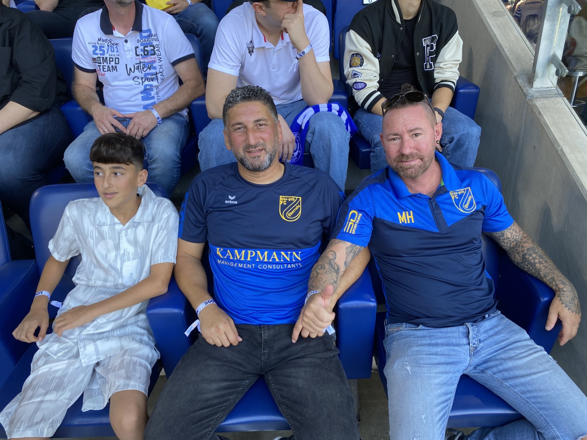 [{"clang_id":1,"value":"Sechs Männer und ein Junge sitzen auf blauen Stadion-Sitzen. Sie tragen blaue und weiße Kleidung, einige mit Sportverein-Logos."}]