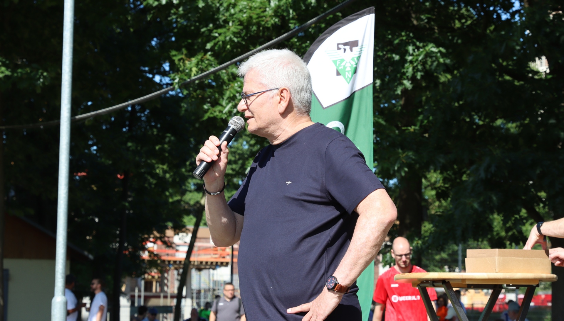 [{"clang_id":1,"value":"Mann mit Mikrofon spricht vor Publikum im Freien. Im Hintergrund grüne Bäume und eine Fahne mit Wappen."}]
