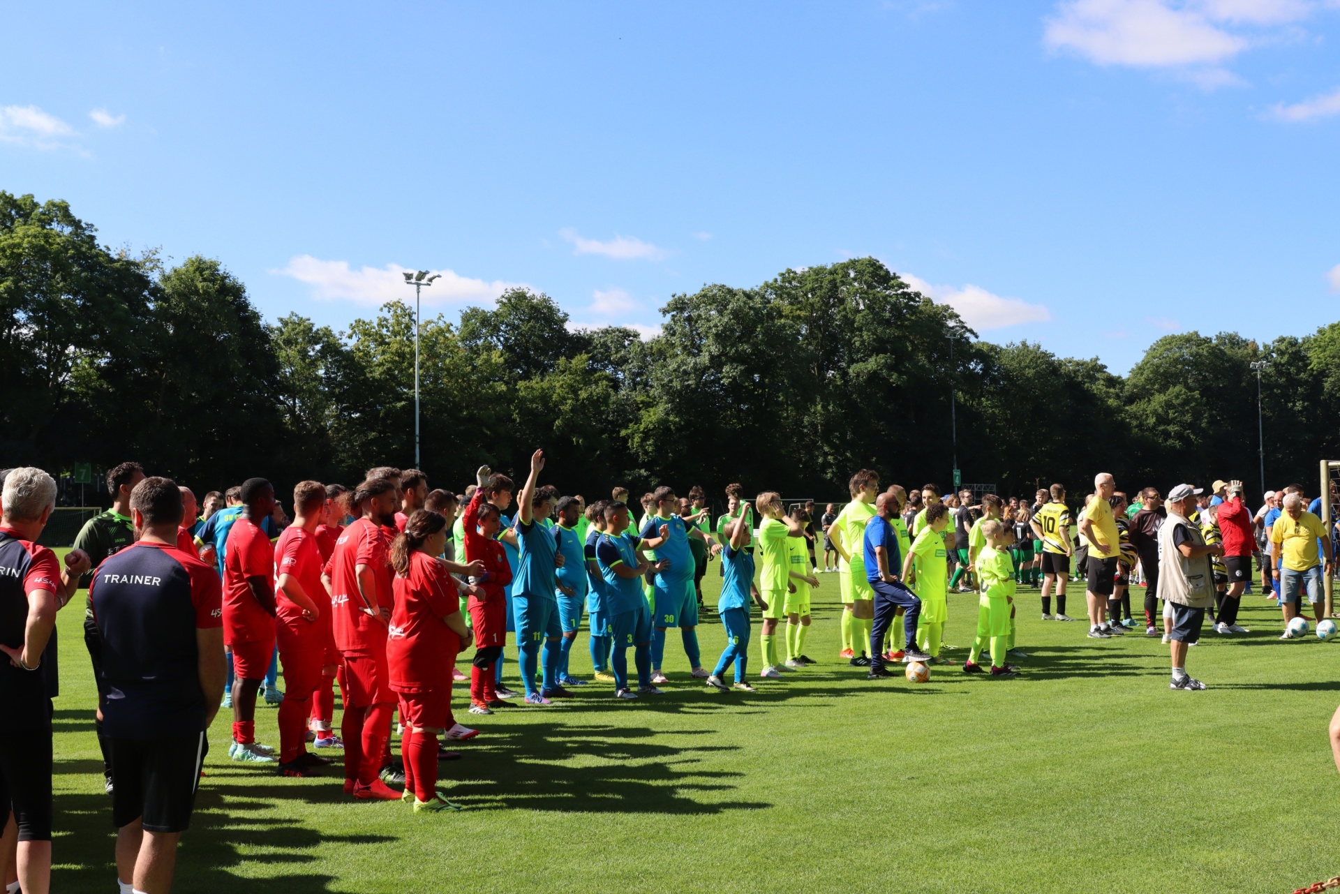 [{"clang_id":1,"value":"Fußballspiel mit zwei Mannschaften in roten und blauen/hellgrünen Trikots. Zuschauer stehen am Spielfeldrand unter blauem Himmel mit Bäumen im Hintergrund."}]