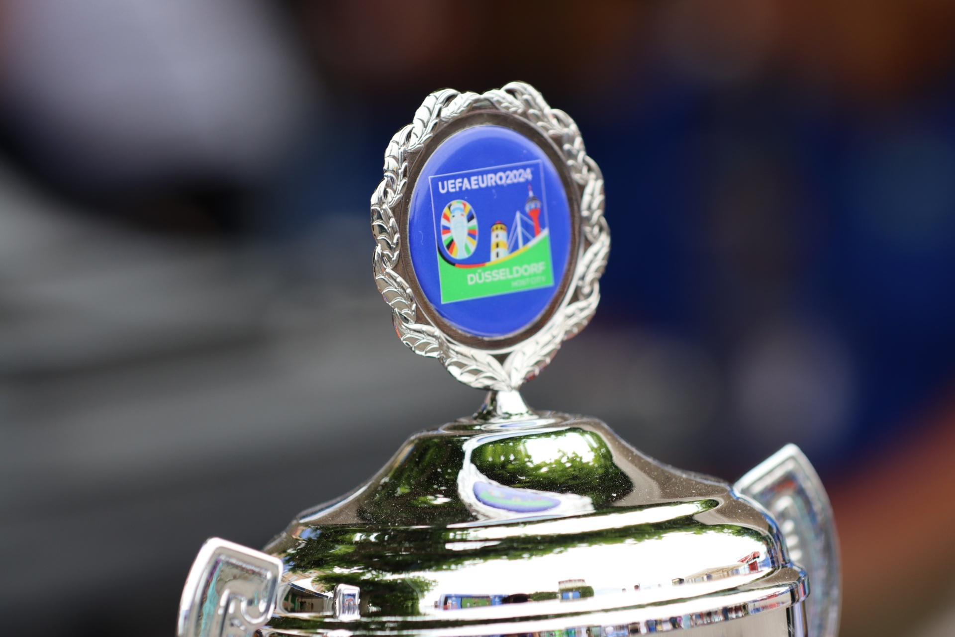 [{"clang_id":1,"value":"Pokal mit UEFA EURO 2024 Logo und dem Schriftzug \"DÜSSELDORF\". Silberner Pokal mit blauer und grüner Dekoration. Im Hintergrund verschwommene Stadiontribünen."}]