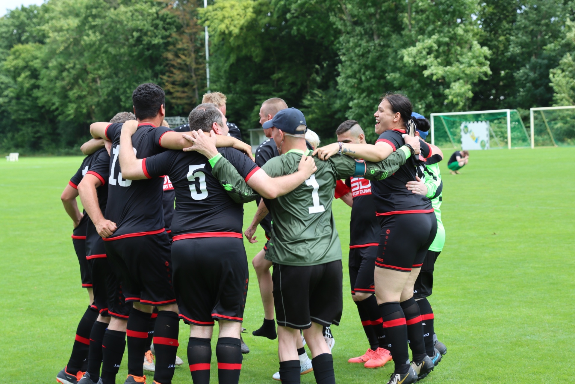 [{"clang_id":1,"value":"Fußballteam umarmt sich jubelnd auf grünem Rasen. Spieler tragen schwarze Trikots mit roten Akzenten. Im Hintergrund Bäume und Fußballtor."}]
