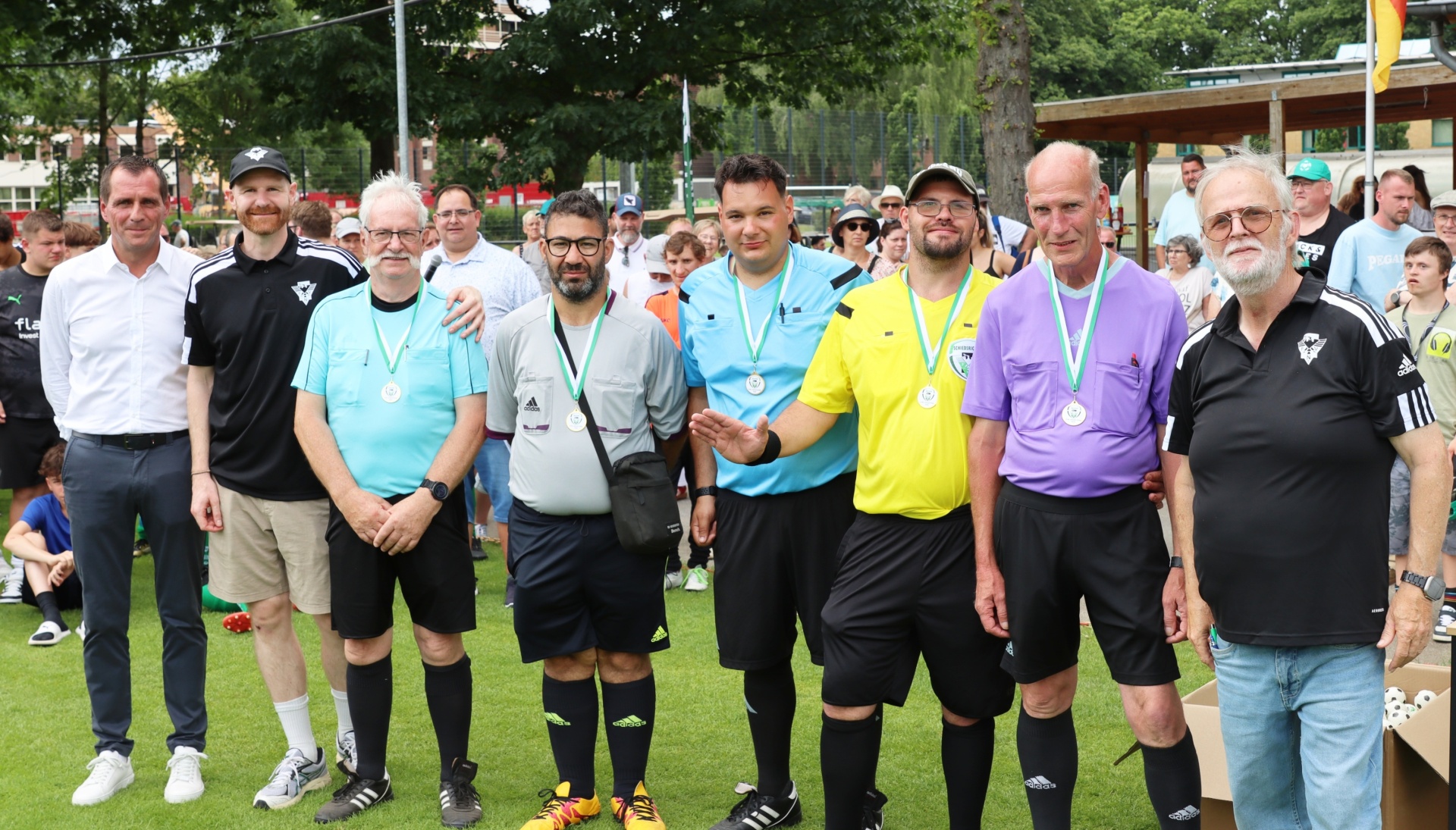[{"clang_id":1,"value":"Gruppe von Männern in Sportkleidung und Schiedsrichter-Outfit auf Grünfläche, einige mit Pfeifen um den Hals. Im Hintergrund Personen und Gebäude."}]