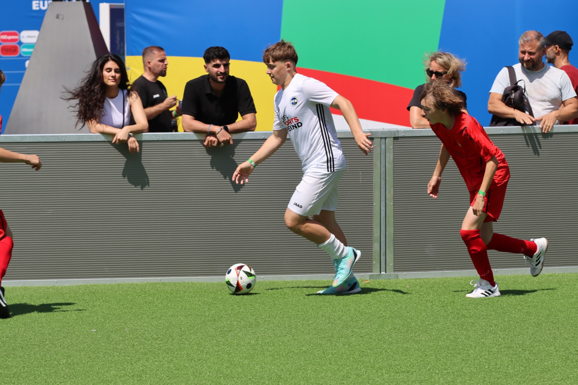 [{"clang_id":1,"value":"Zwei Fußballspieler auf grünem Rasen, einer in rot, der andere in weißem Trikot. Im Hintergrund eine Zuschauertribüne mit Menschen. Bunte Wände im Hintergrund."}]