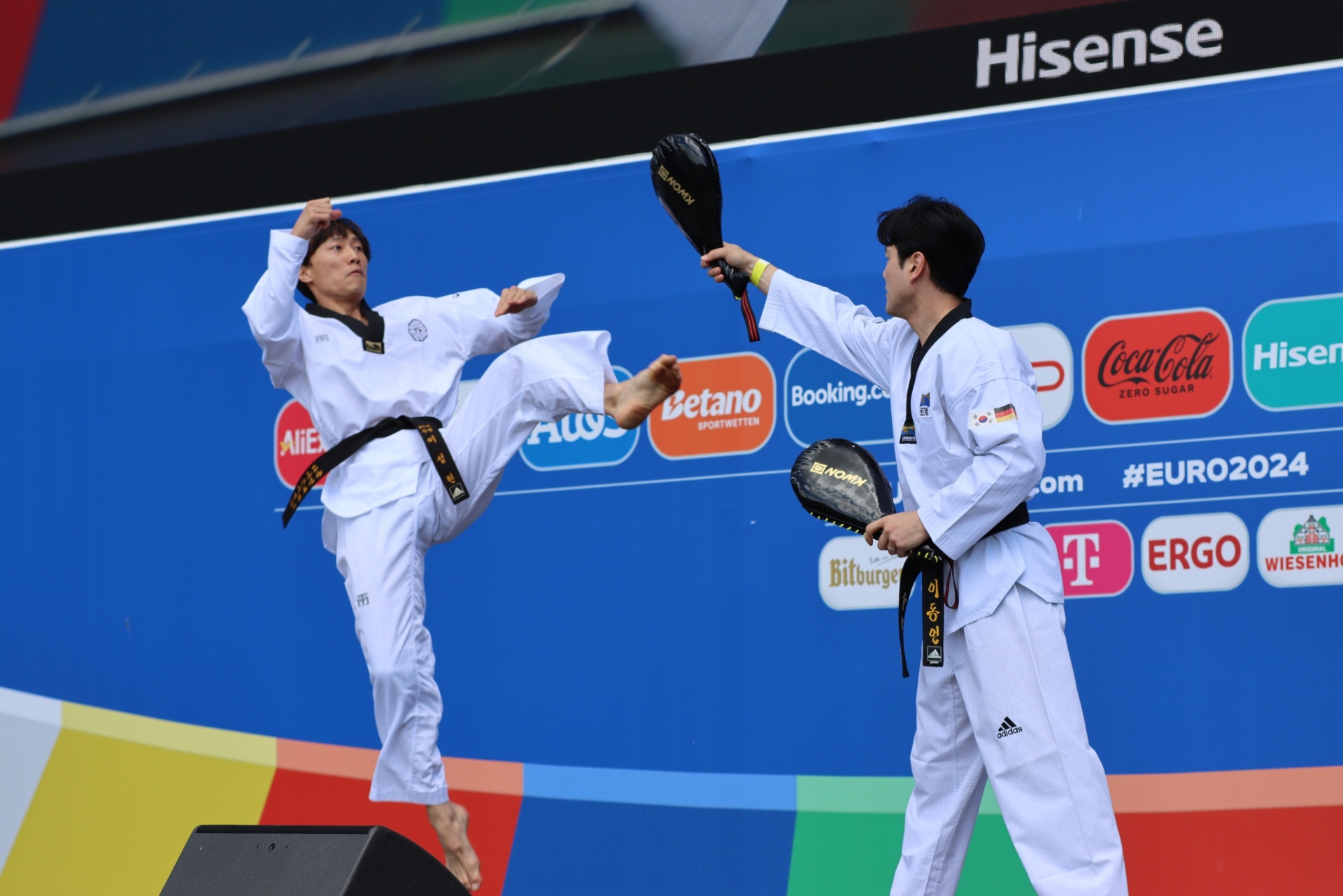 [{"clang_id":1,"value":"Zwei Taekwondo-Kämpfer in weißen Uniformen treten auf einer blauen Matte vor einem Werbebanner gegeneinander an. Einer springt und führt einen Tritt aus."}]