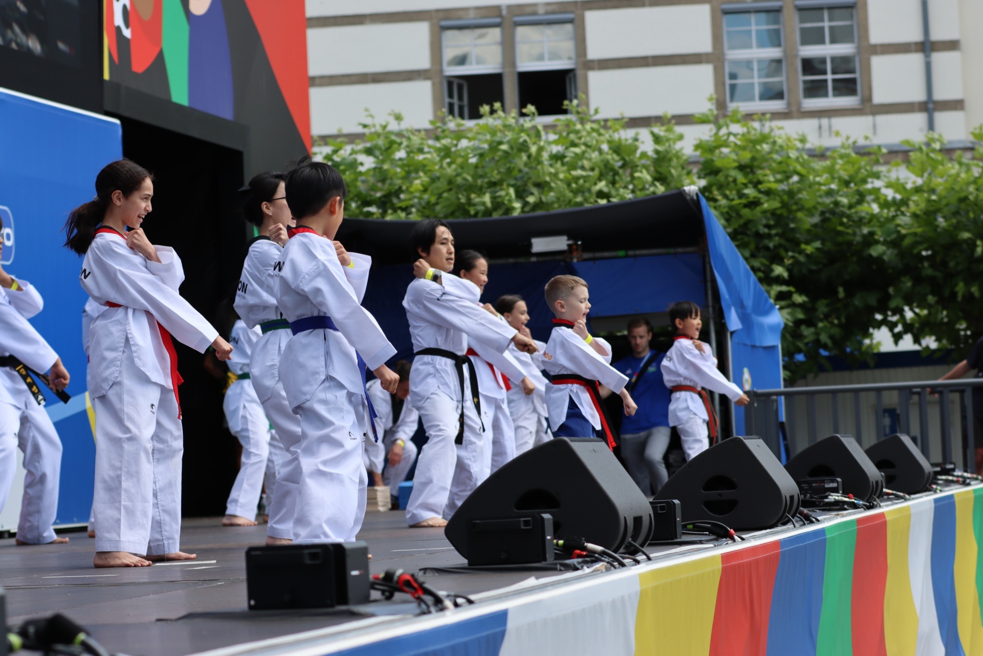 [{"clang_id":1,"value":"Taekwondo-Gruppe auf erhöhter Bühne. Kinder und Erwachsene in weißen Anzügen mit schwarzen Gürteln führen eine Bewegung aus. Hintergrund: Gebäude und Grünflächen."}]