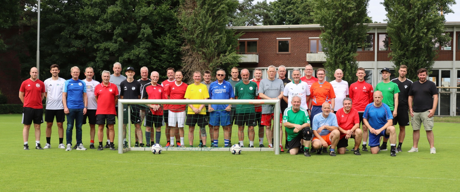 [{"clang_id":1,"value":"Fußballmannschaften in farbigen Trikots stehen auf einem Rasenplatz vor einem Fußballtor und einem Gebäude mit Bäumen im Hintergrund."}]