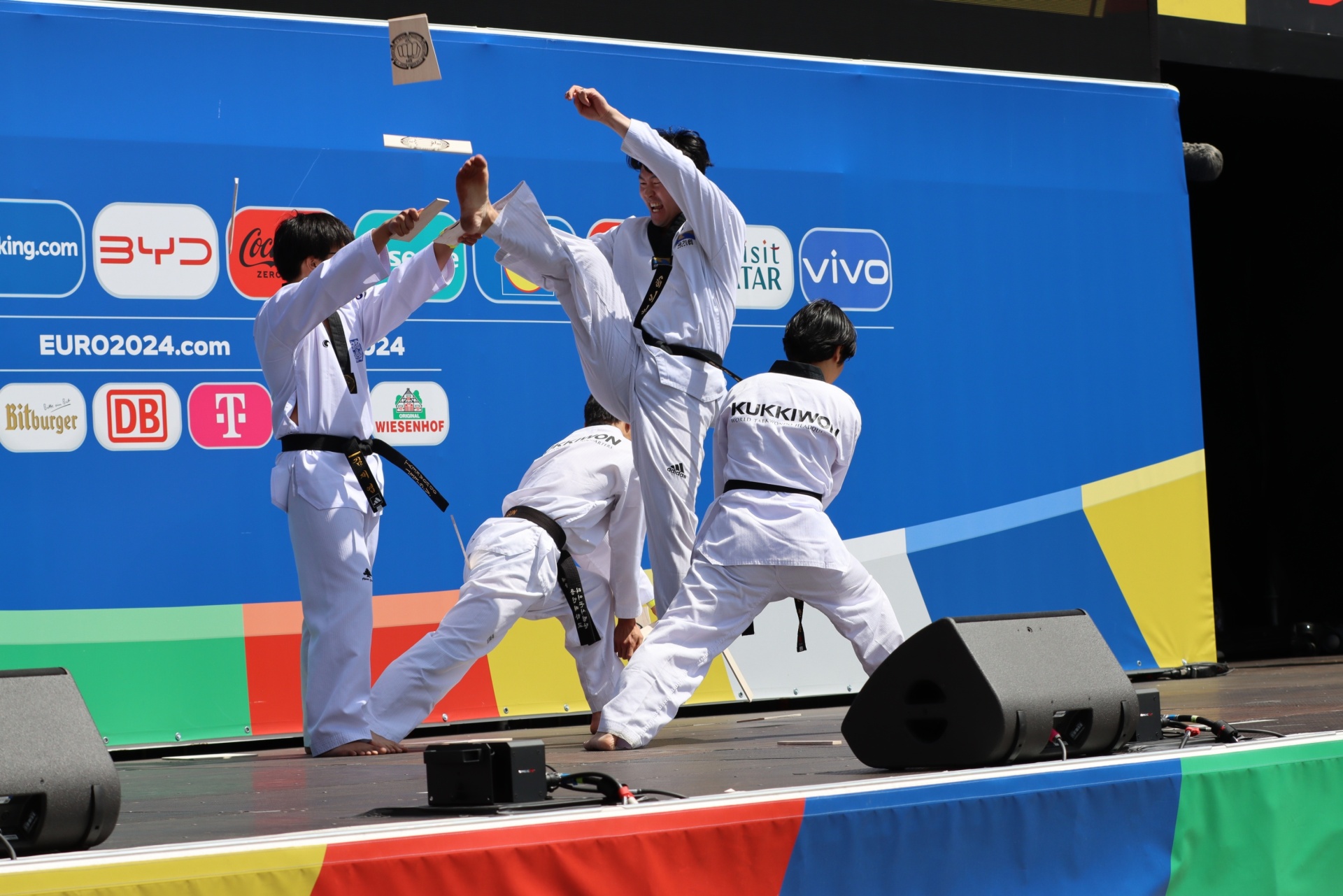 [{"clang_id":1,"value":"Vier Taekwondo-Sportler in weißen Anzügen führen eine synchronisierte Bewegung auf einer Bühne vor blauem Werbebanner auf."}]