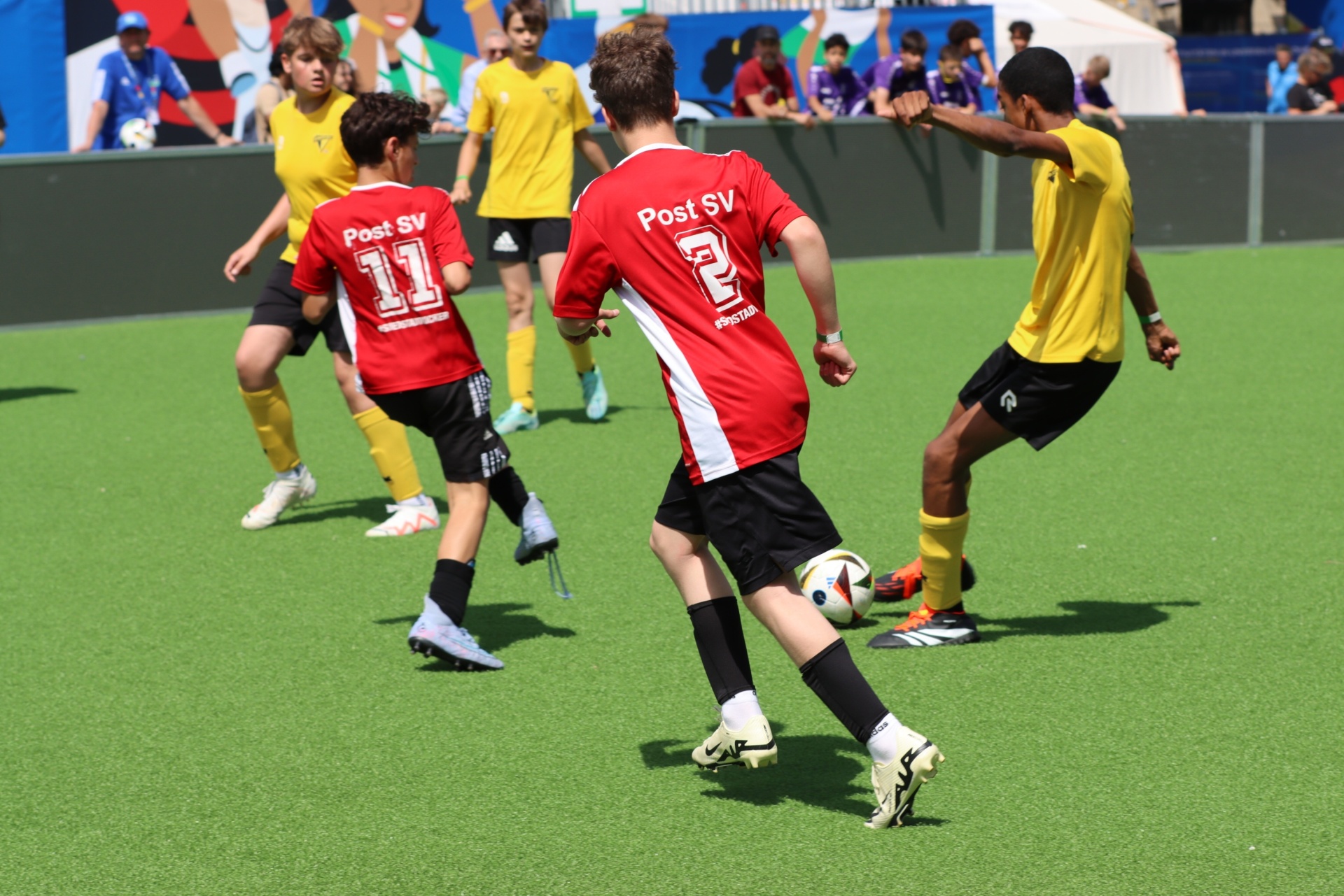 [{"clang_id":1,"value":"Fußballspiel: Zwei Mannschaften mit roten und gelben Trikots auf grünem Kunstrasen. Spieler mit Nummern 2 und 11 im Vordergrund. Zuschauer im Hintergrund."}]