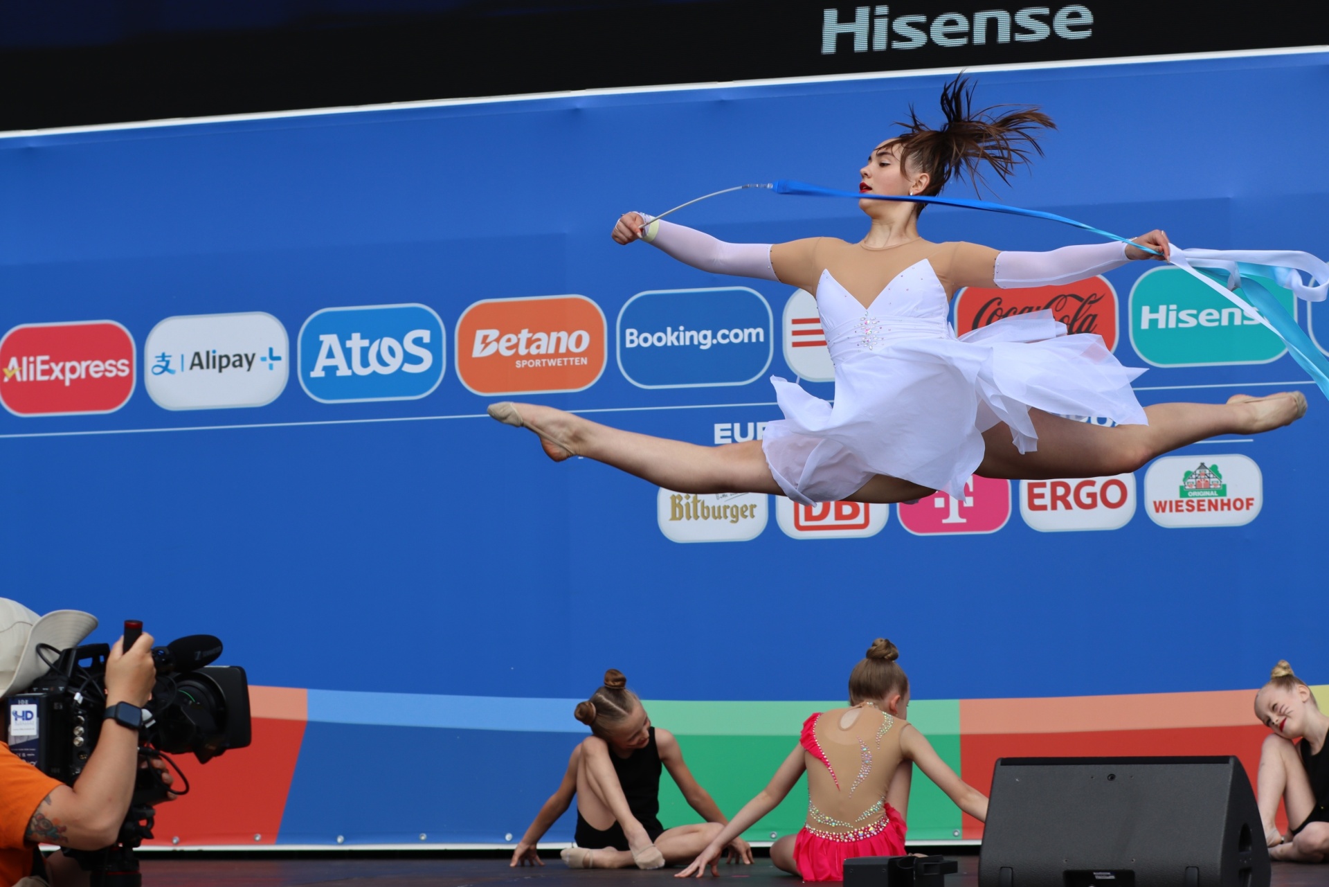 [{"clang_id":1,"value":"Rhythmische Sportgymnastin in weißem Outfit springt mit blauem Band. Im Hintergrund weitere Gymnastinnen und Werbebanner."}]