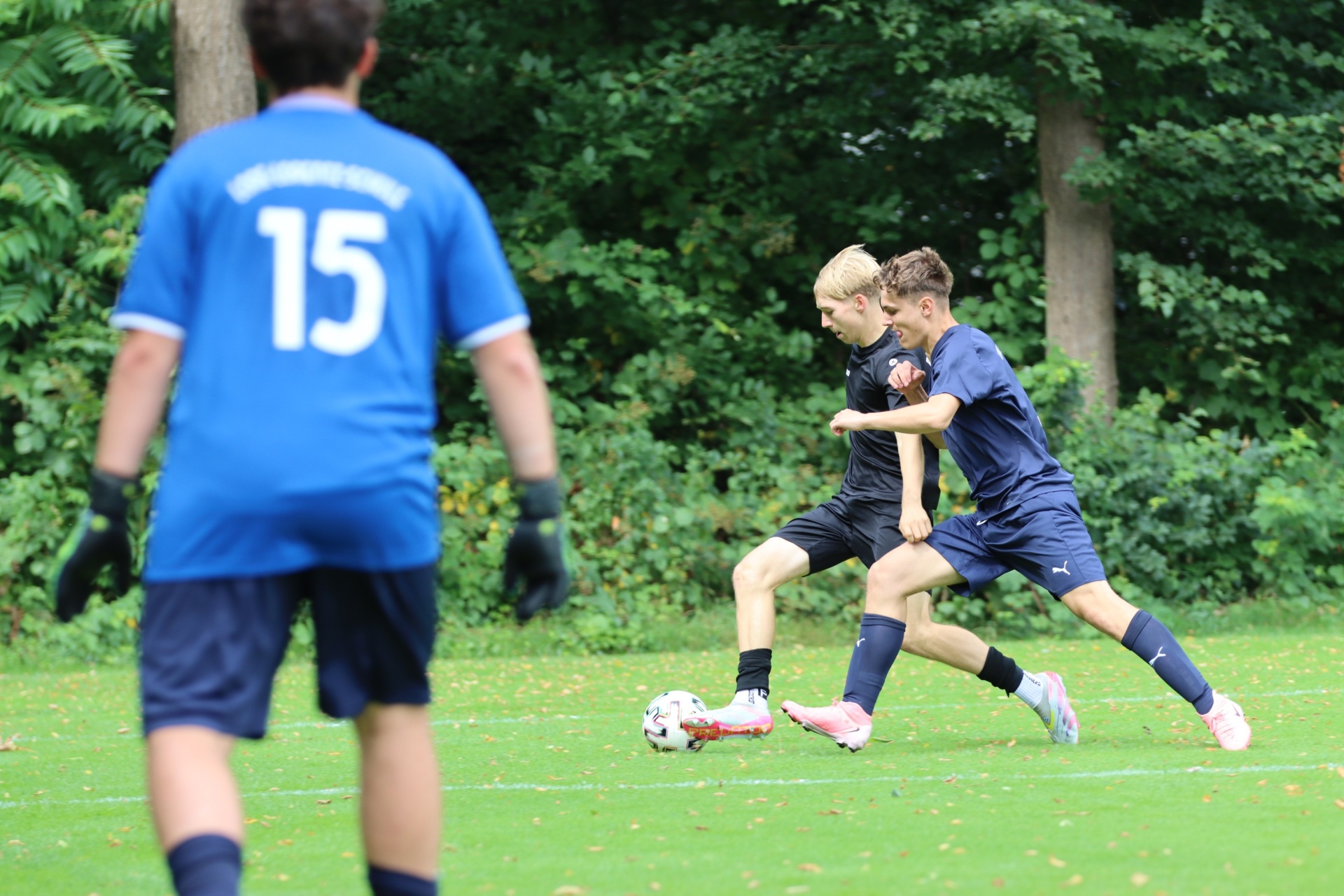 [{"clang_id":1,"value":"Zwei Fussballspieler kämpfen um den Ball auf grünem Rasen. Im Hintergrund steht ein Spieler mit der Nummer 15 auf dem Trikot und Bäume."}]