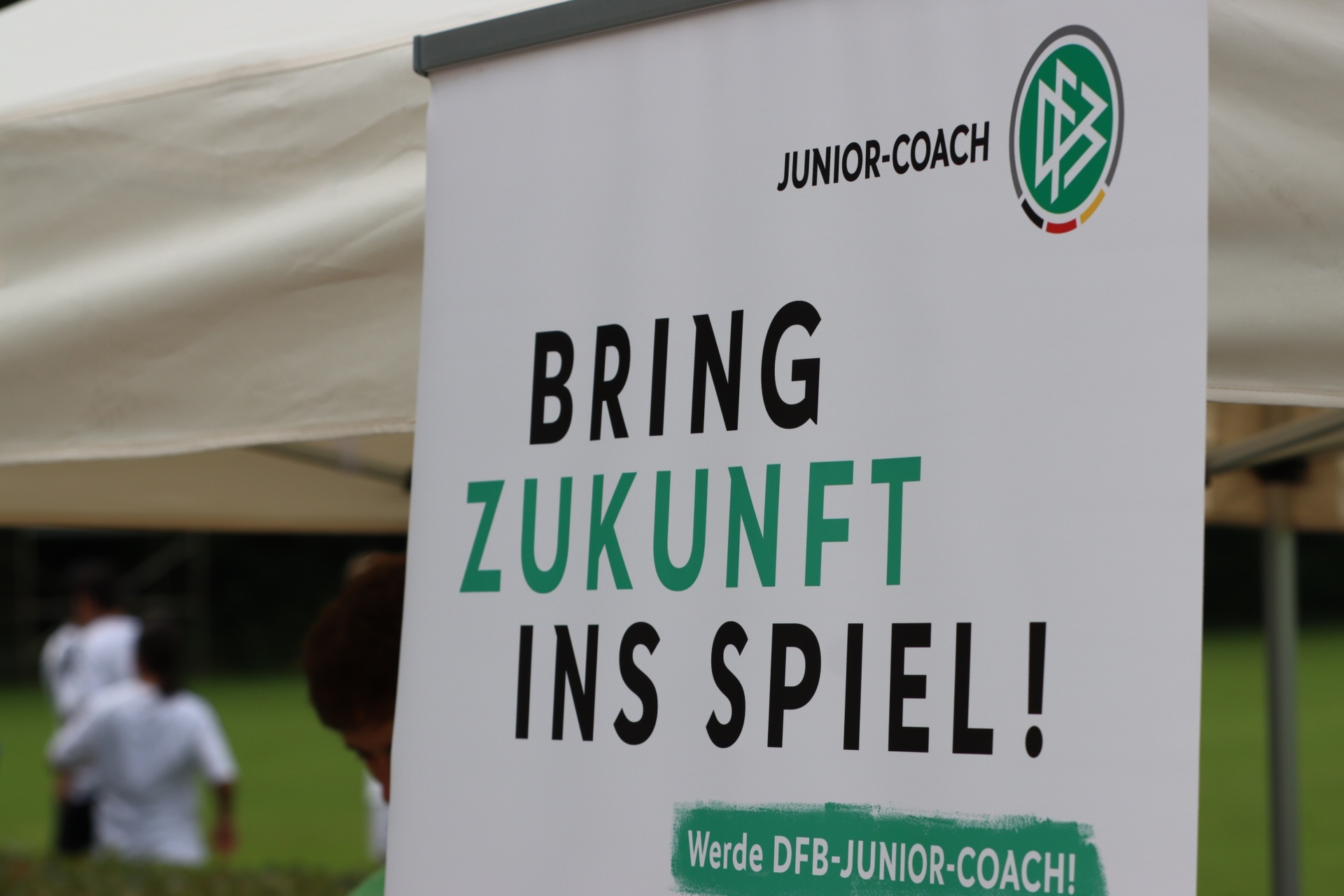 [{"clang_id":1,"value":"Banner mit Aufschrift \"BRING ZUKUNFT INS SPIEL!\", \"JUNIOR-COACH\" und DFB-Logo. Im Hintergrund verschwommen Fußballspieler auf grünem Rasen unter einem Zelt."}]