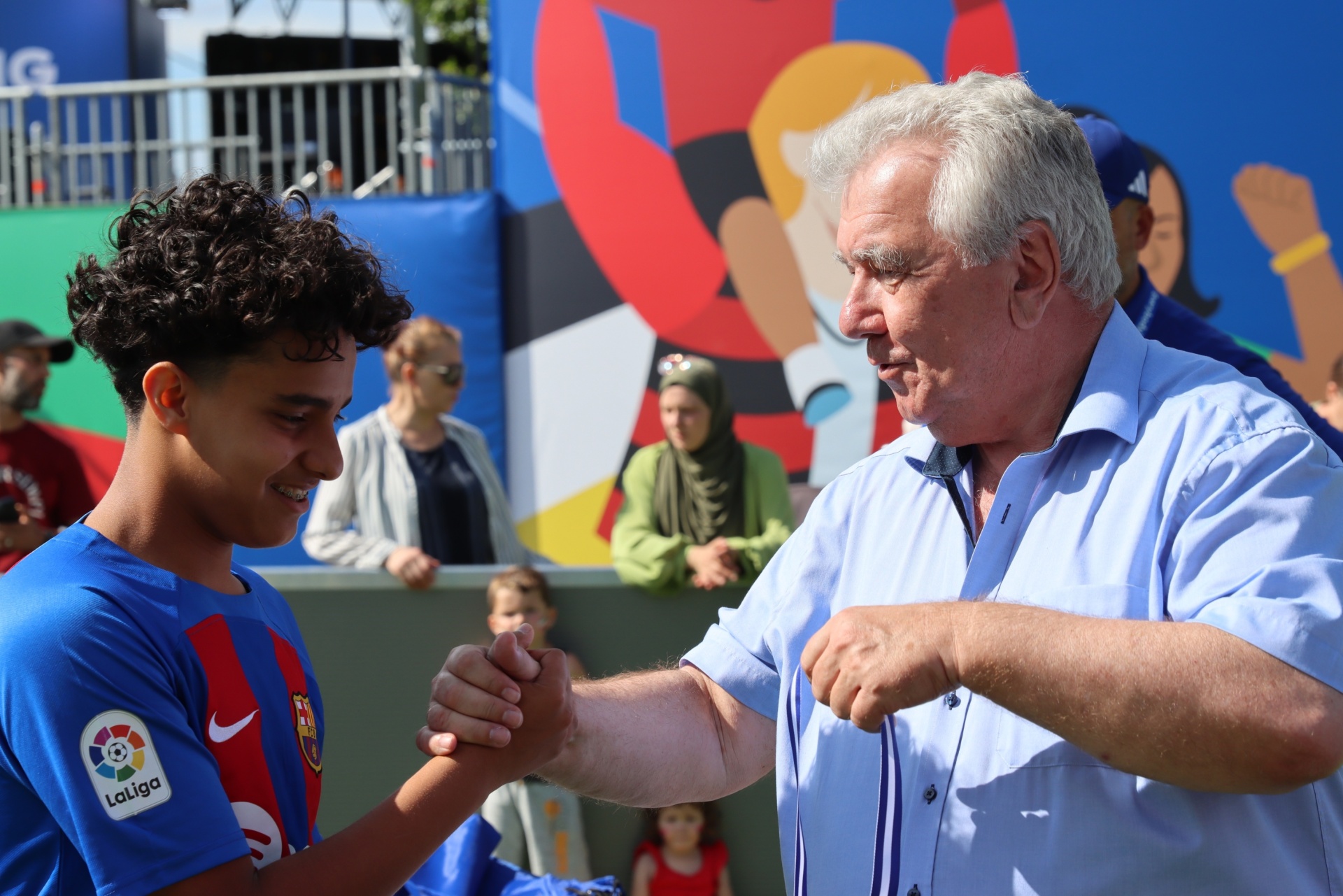 [{"clang_id":1,"value":"Zwei Männer, einer mit lockigem Haar im FC Barcelona Trikot, geben sich die Hände. Im Hintergrund eine Menge Menschen und bunte Dekoration."}]