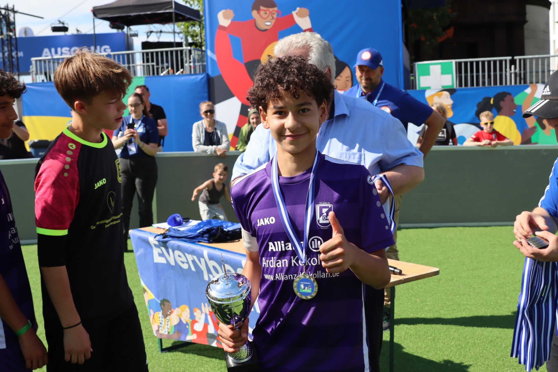[{"clang_id":1,"value":"Junge hält Pokal, flankiert von zwei weiteren Jungen. Im Hintergrund Menschen und Banner bei einer Sportveranstaltung."}]