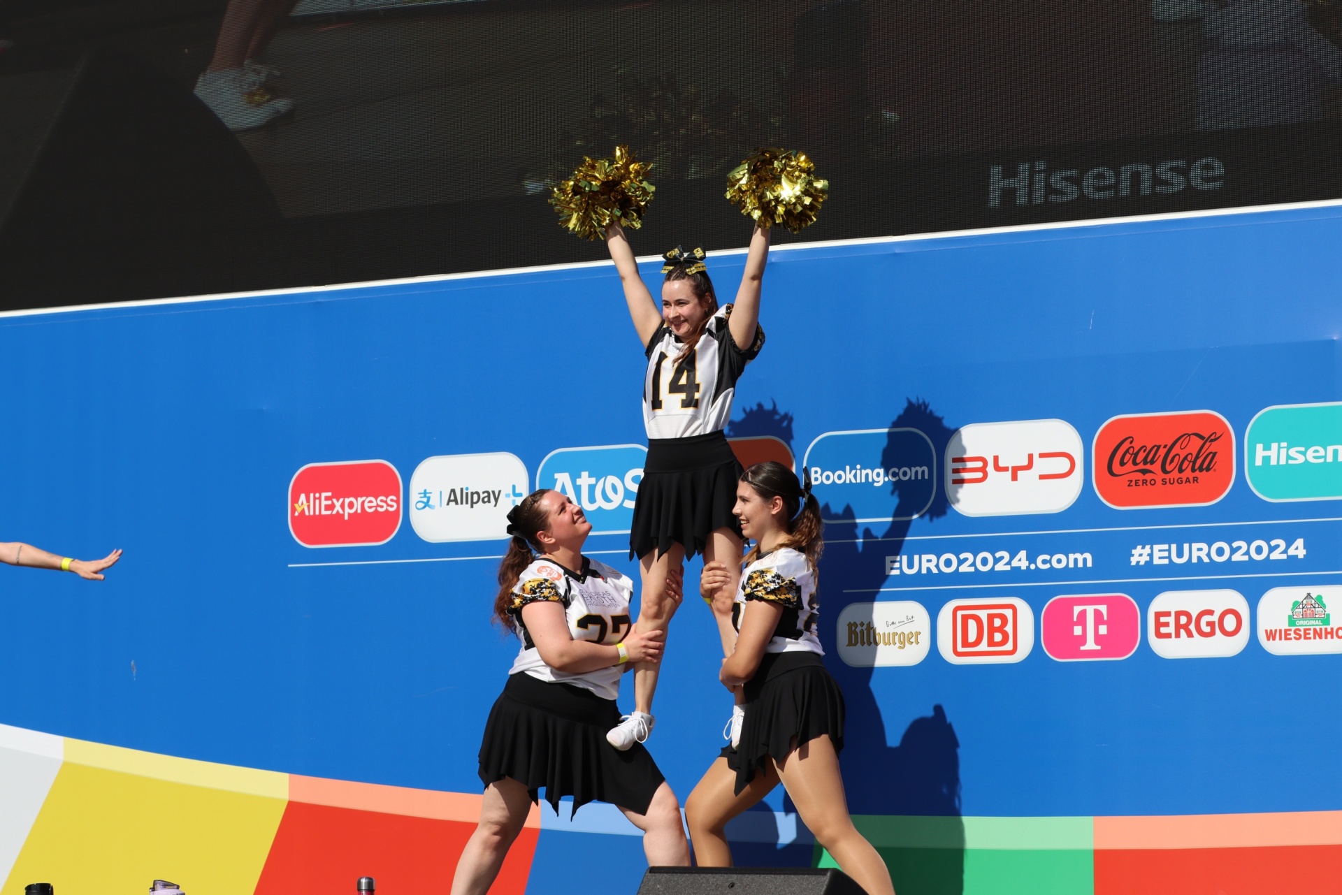 [{"clang_id":1,"value":"Cheerleader-Pyramide vor blauem Werbebanner mit Sponsorenlogos (AliExpress, Coca-Cola, Hisense u.a.). Eine Cheerleaderin wird hochgehoben, die anderen bilden die Basis."}]