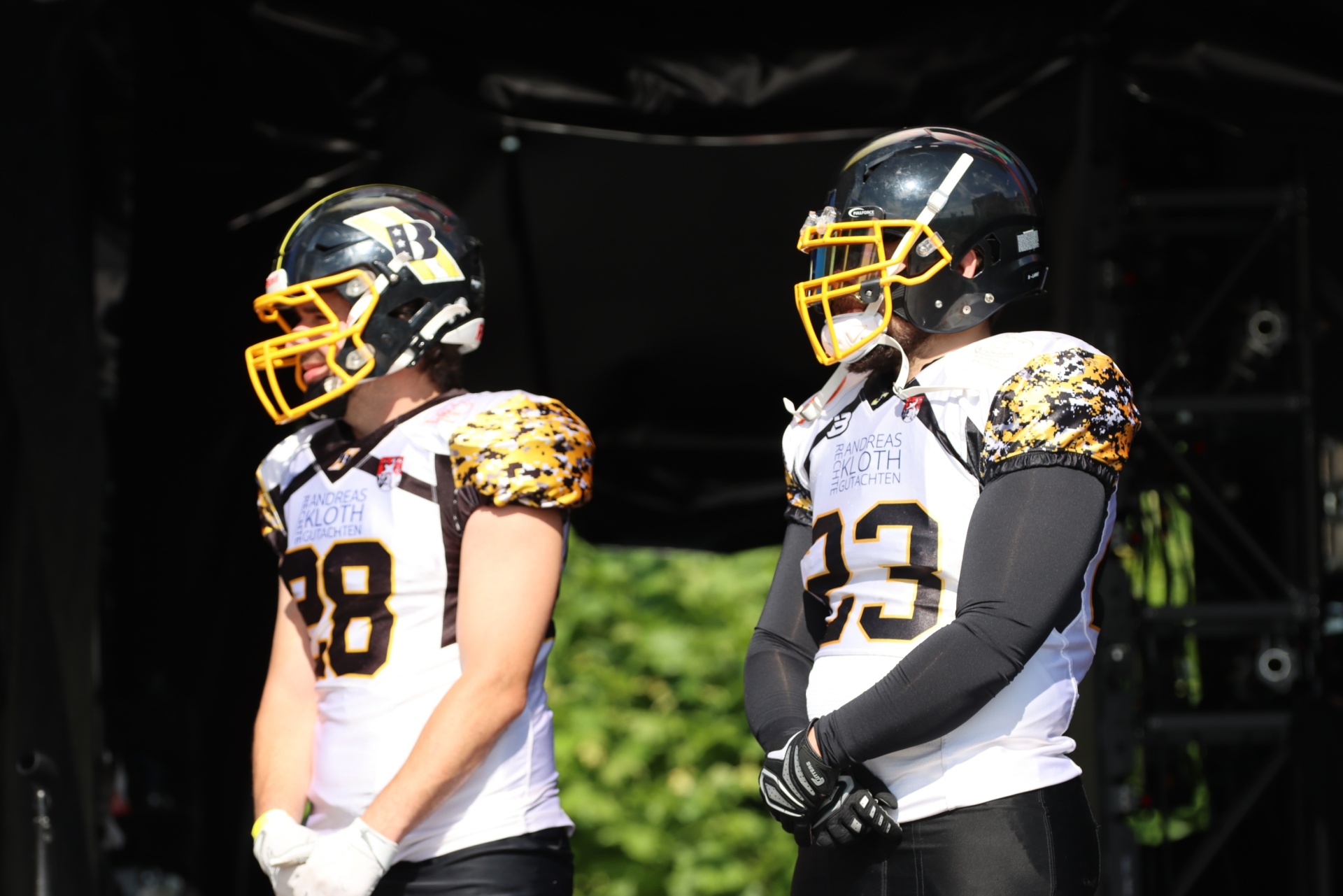 [{"clang_id":1,"value":"Zwei American-Football-Spieler in weißen Trikots mit Nummer 13 und 98, schwarzer Hose und Helmen stehen nebeneinander auf einem Spielfeld."}]