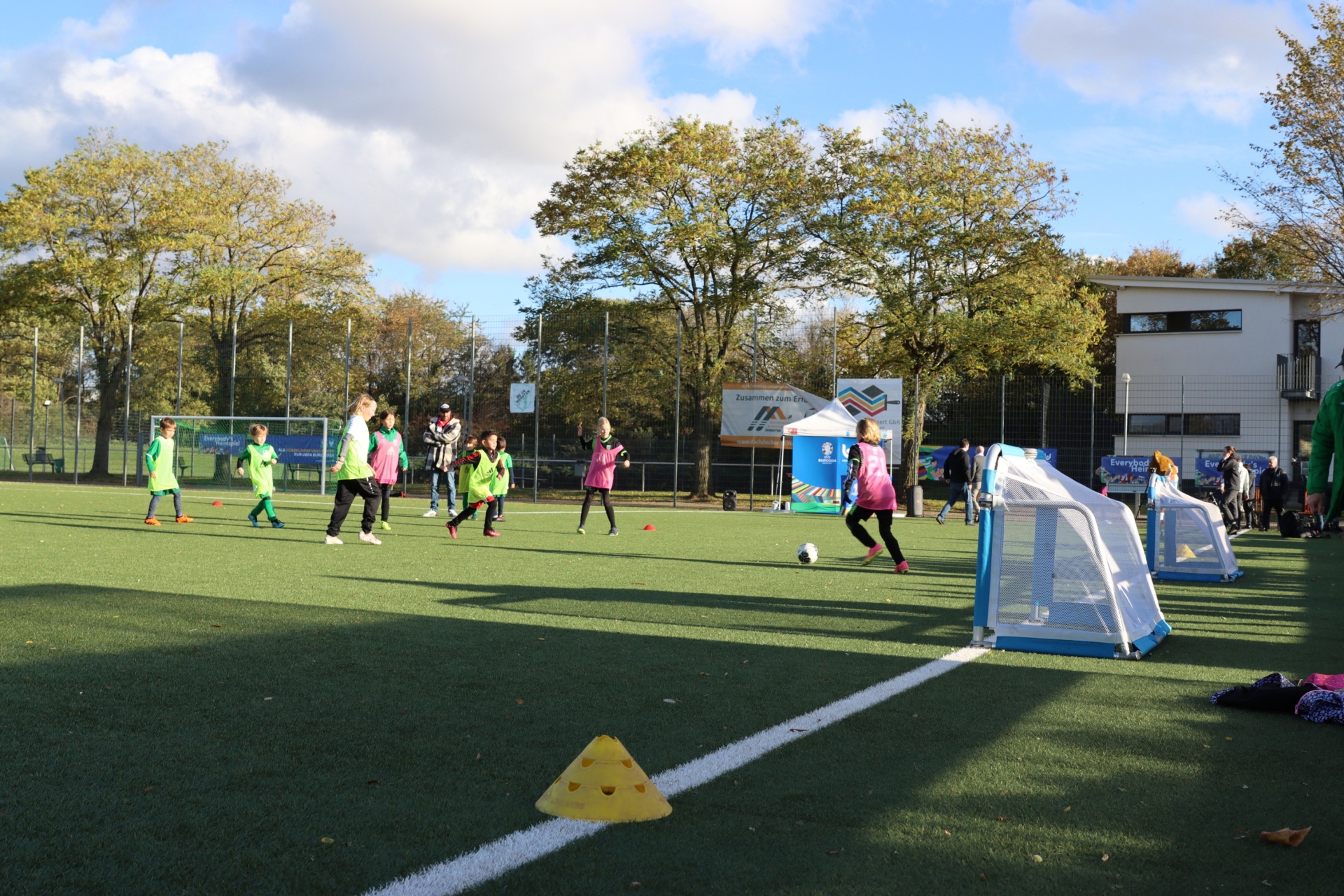 [{"clang_id":1,"value":"Fußballtraining mit Kindern auf grünem Kunstrasenplatz. Im Hintergrund Bäume und ein Gebäude. Einige Spieler tragen bunte Trikots."}]