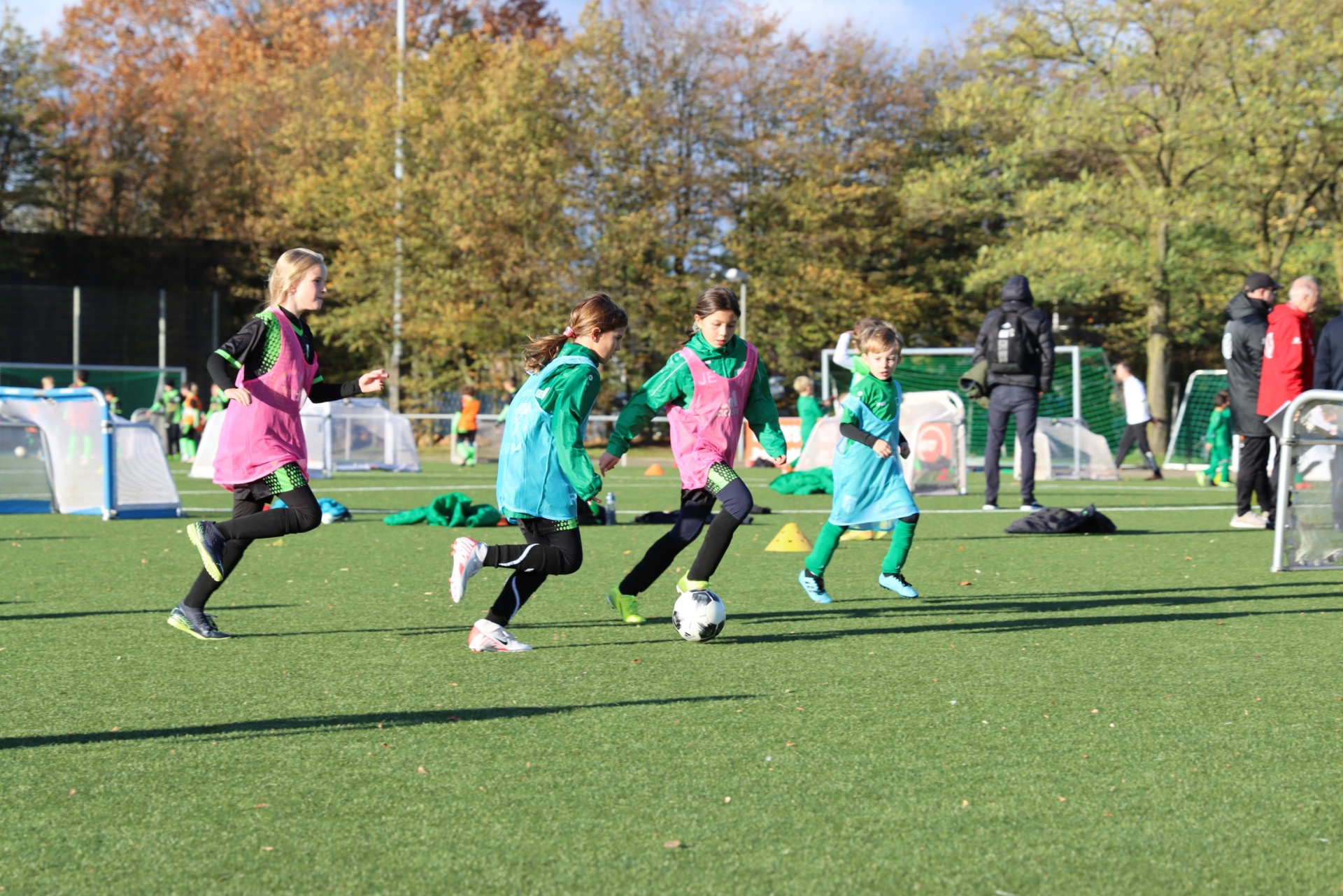 [{"clang_id":1,"value":"Fünf Kinder spielen Fußball auf einem grünen Kunstrasenplatz. Sie tragen Trainingsanzüge in verschiedenen Farben. Im Hintergrund sind Bäume und weitere Personen zu sehen."}]