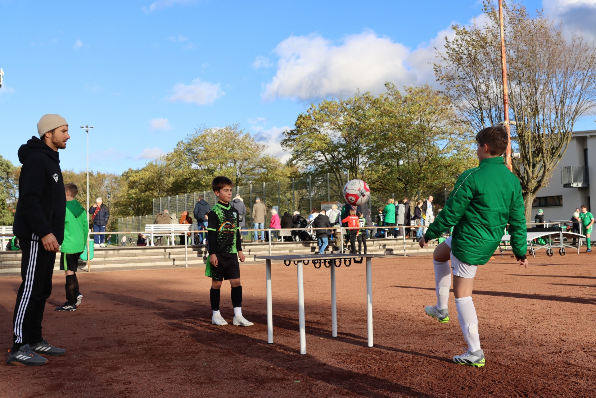 [{"clang_id":1,"value":"Fußballtraining: Trainer und zwei jugendliche Spieler an Hürden auf rotem Tartanplatz. Im Hintergrund weitere Spieler und Zuschauer."}]