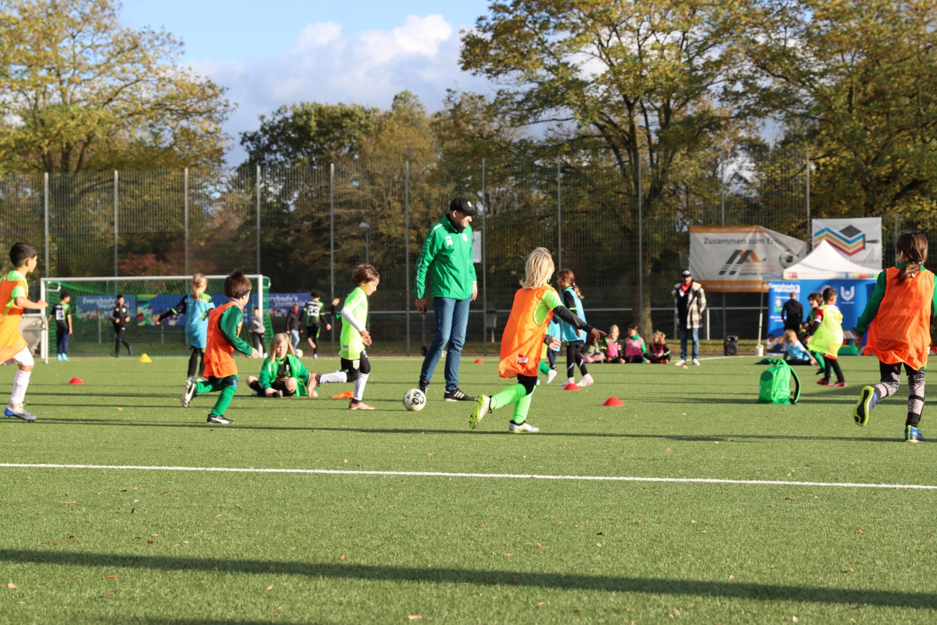 [{"clang_id":1,"value":"Fußballtraining mit Kindern auf Kunstrasenplatz. Trainer und Spieler mit grünen, orangefarbenen oder schwarzen Trainingsanzügen. Bäume im Hintergrund."}]
