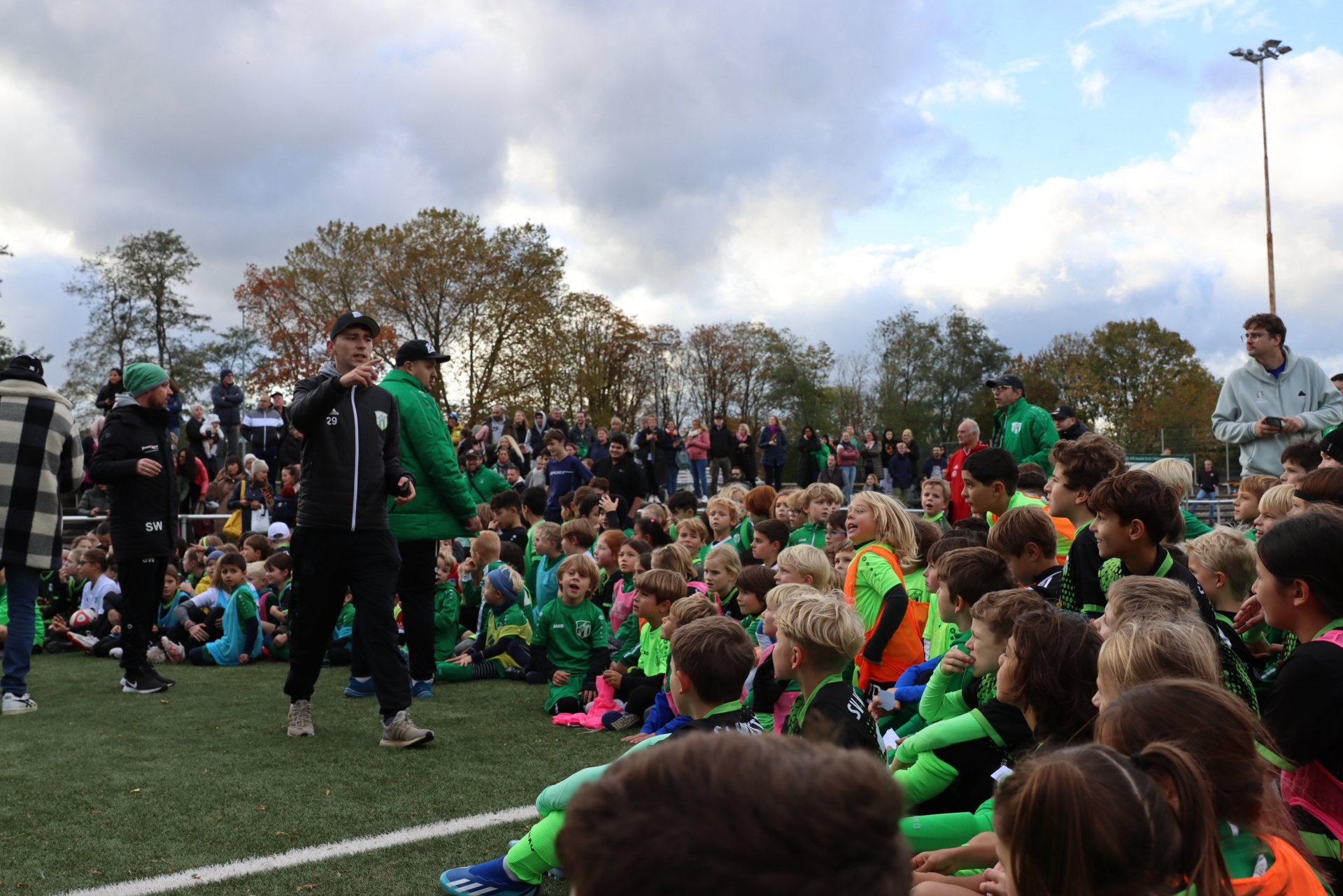 [{"clang_id":1,"value":"Fußballfeld mit vielen Kindern in grün-gelben Trainingsanzügen, die einem Mann mit Mikrofon zuhören. Im Hintergrund Zuschauer und Bäume unter bewölktem Himmel."}]