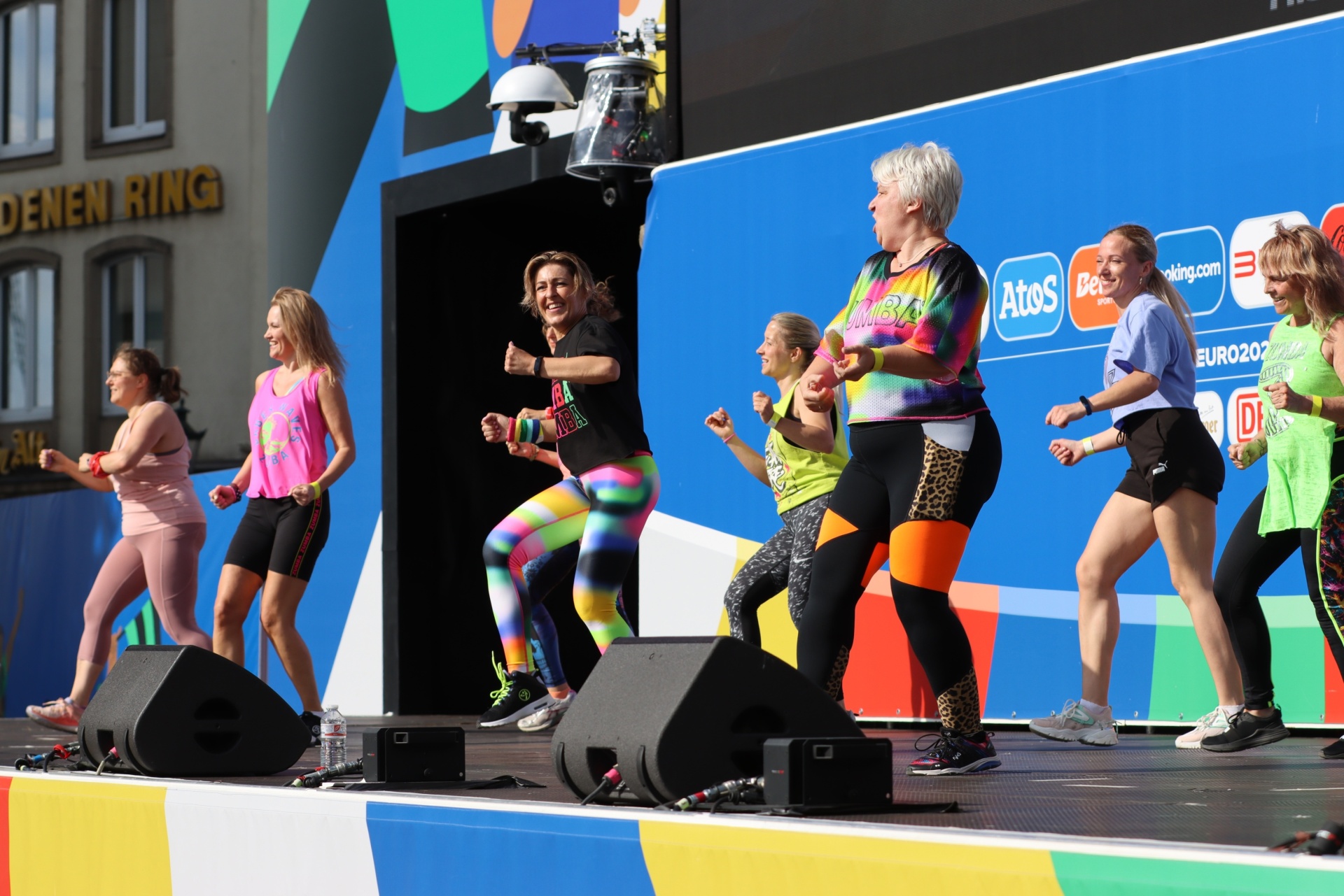 [{"clang_id":1,"value":"Zumba-Kurs auf erhöhter Bühne im Freien. Mehrere Frauen tanzen synchron, bunte Sportkleidung. Im Hintergrund Werbebanner und Publikum."}]