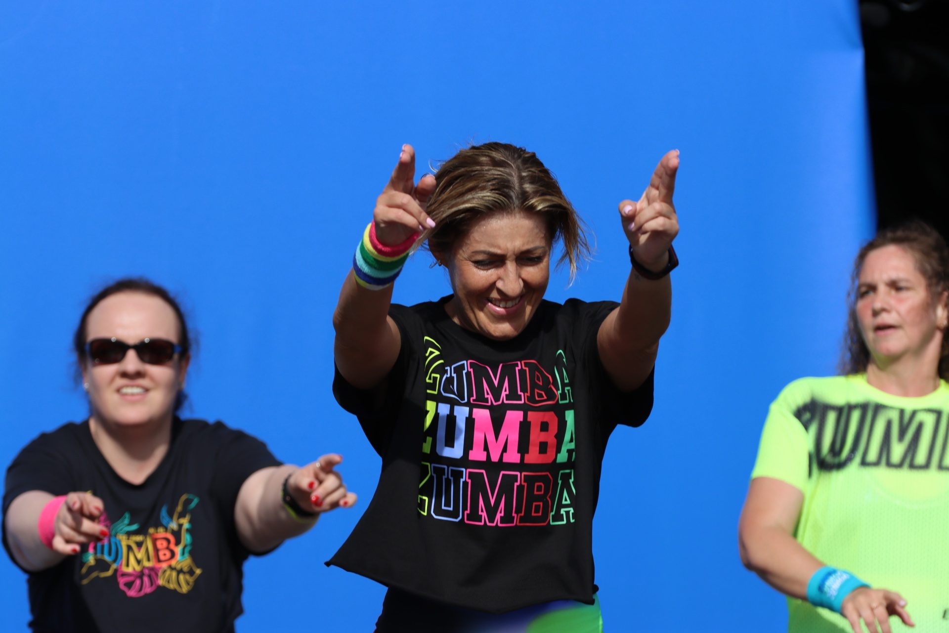 [{"clang_id":1,"value":"Drei Frauen tanzen Zumba vor blauem Hintergrund. Die mittlere Frau trägt ein schwarzes T-Shirt mit \"ZUMBA\" Aufdruck und hebt die Hände."}]