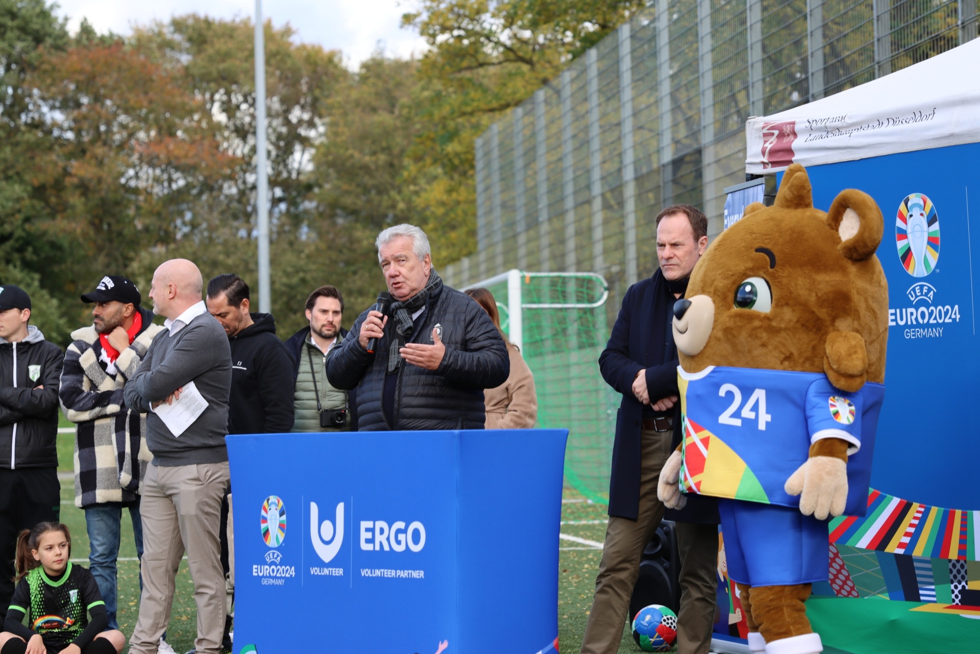 [{"clang_id":1,"value":"Mann am Rednerpult hält Rede vor Publikum und großem blauem Maskottchen mit \"24\" auf dem Bauch. Im Hintergrund Sportplatz und Werbebanner für EURO 2024."}]