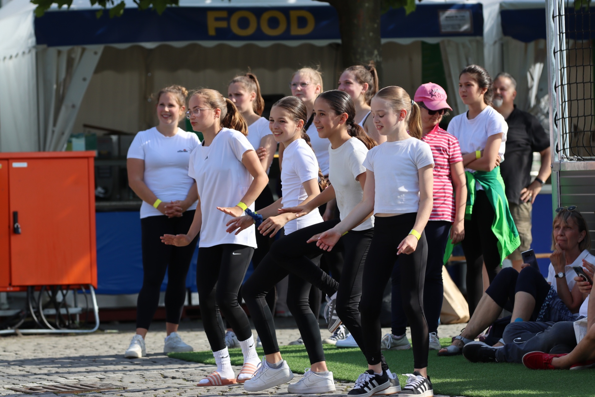 [{"clang_id":1,"value":"Gruppe junger Tänzerinnen in weißen Oberteilen und schwarzen Hosen trainiert im Freien vor einem Essensstand. Zuschauende sitzen auf Bänken."}]