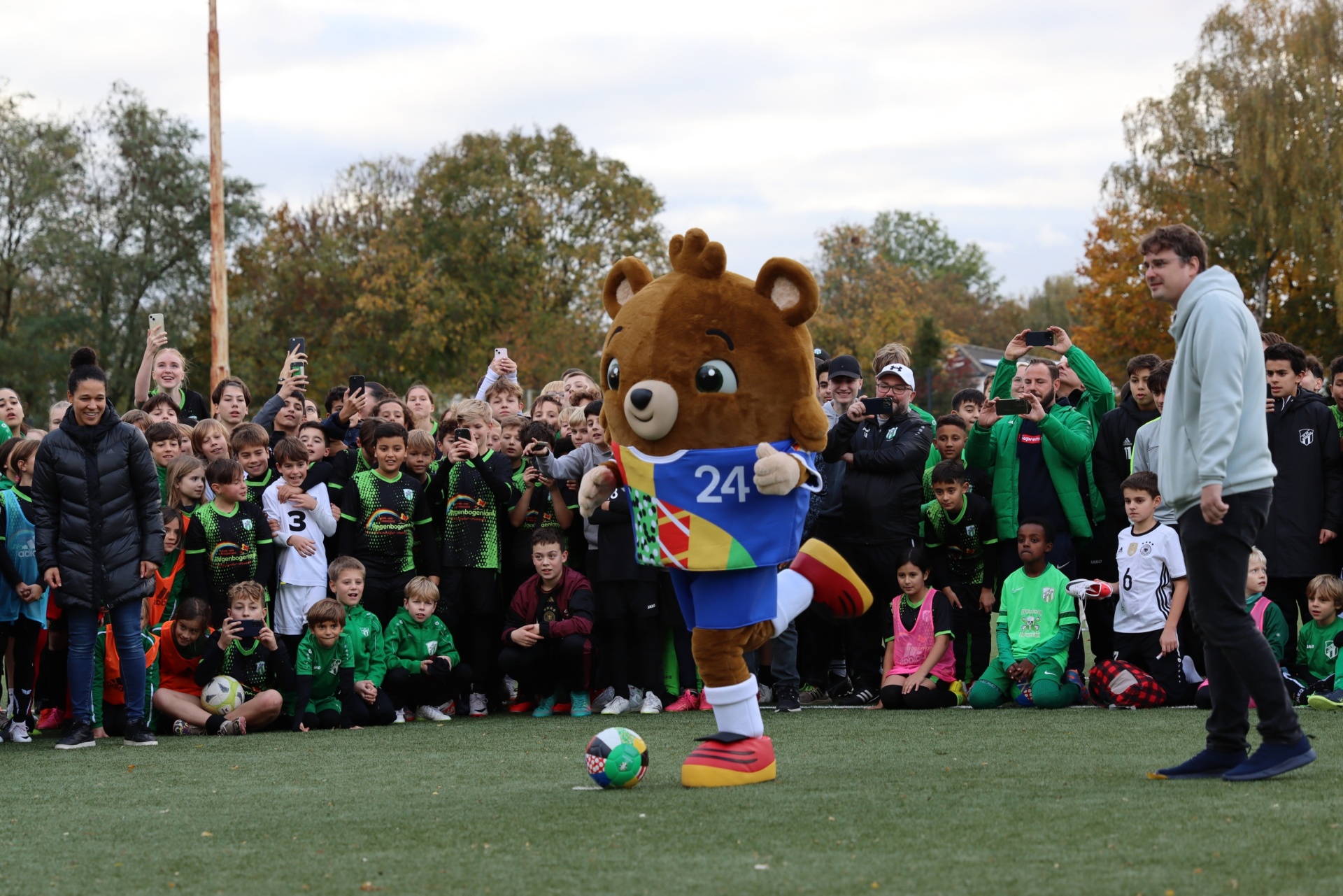 [{"clang_id":1,"value":"Fußballfeld mit Kindern in Sportkleidung. Ein Bär in Fußballtrikot dribbelt einen Ball. Im Hintergrund stehen Erwachsene und Bäume."}]