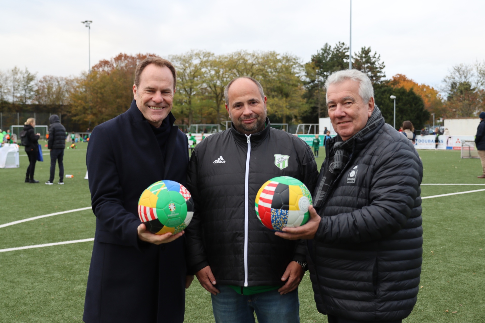 [{"clang_id":1,"value":"Drei Männer stehen auf einem Fußballfeld. Jeder hält einen bunten Fußball. Links ein Mann mit dunklem Mantel, rechts ein Mann mit schwarzer Jacke."}]