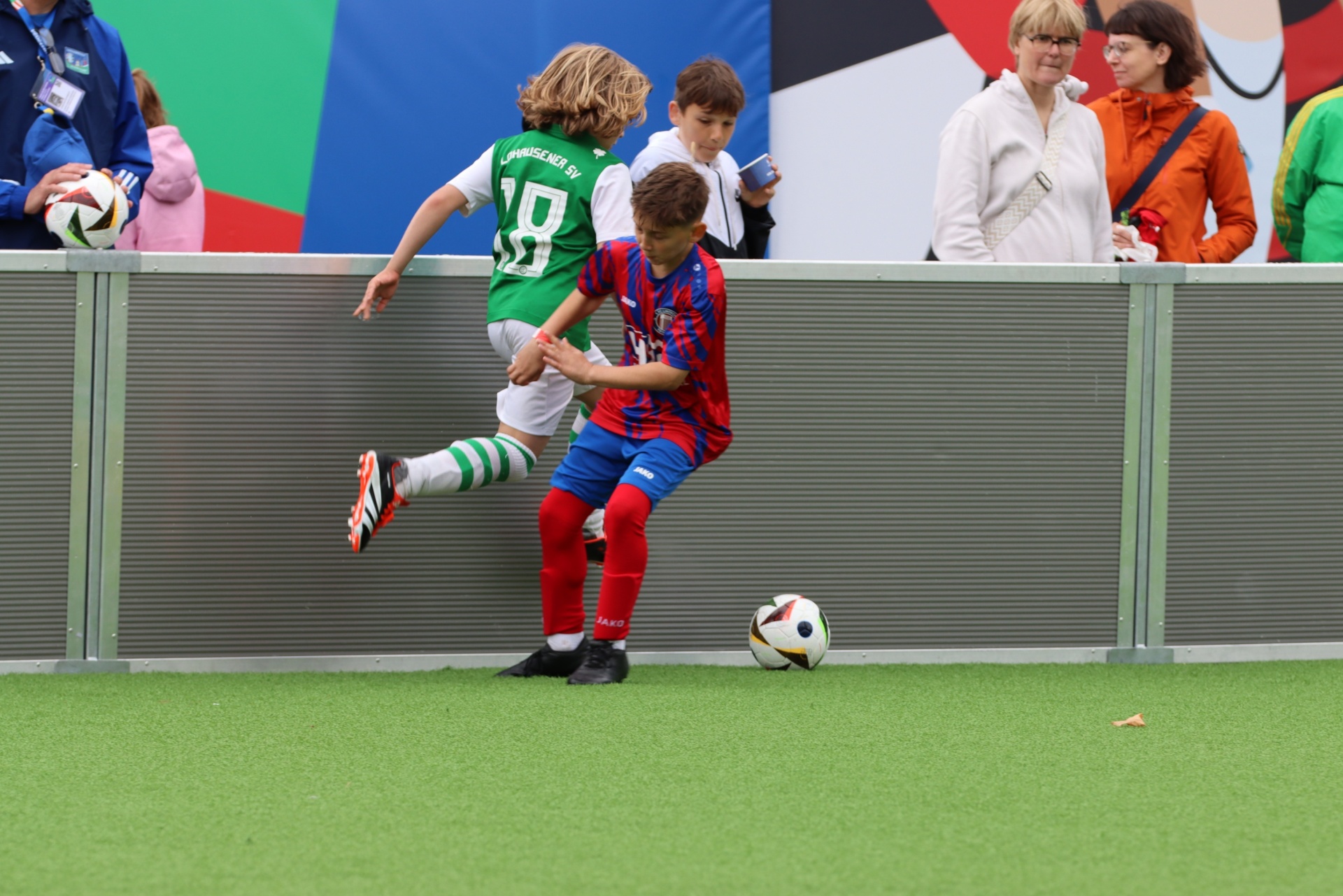 [{"clang_id":1,"value":"Zwei junge Fußballspieler im Spiel. Links grünes Trikot mit Nummer 8, rechts blau-rotes Trikot mit roten Socken. Zuschauer im Hintergrund."}]
