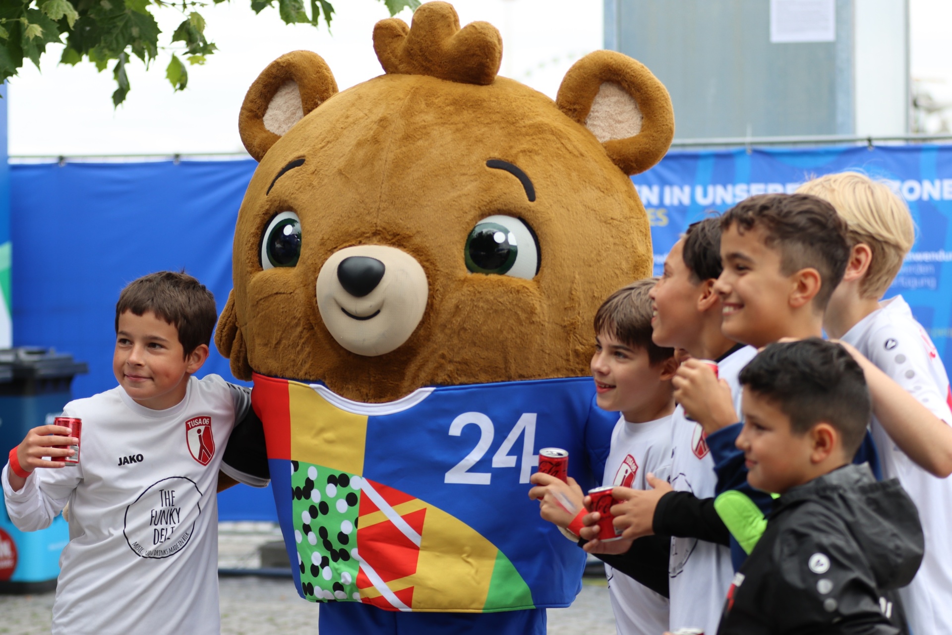 [{"clang_id":1,"value":"Fußballmannschaft mit Maskottchen: Kinder in Fußballtrikots stehen um ein großes Bären-Maskottchen, das ein Trikot mit der Zahl 24 trägt."}]