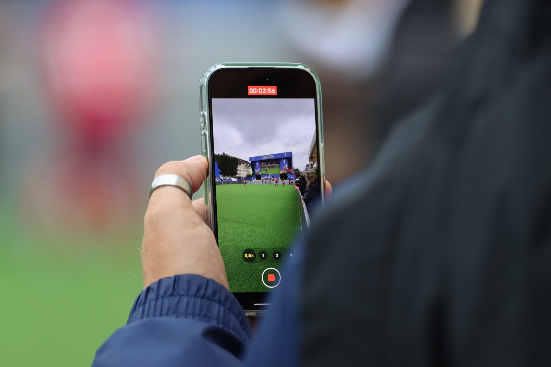 [{"clang_id":1,"value":"Hand hält Smartphone, das ein Fußballfeld filmt. Auf dem Display ist die Zeit 00:02:56 zu sehen. Hintergrund unscharf, Person mit dunkler Jacke."}]