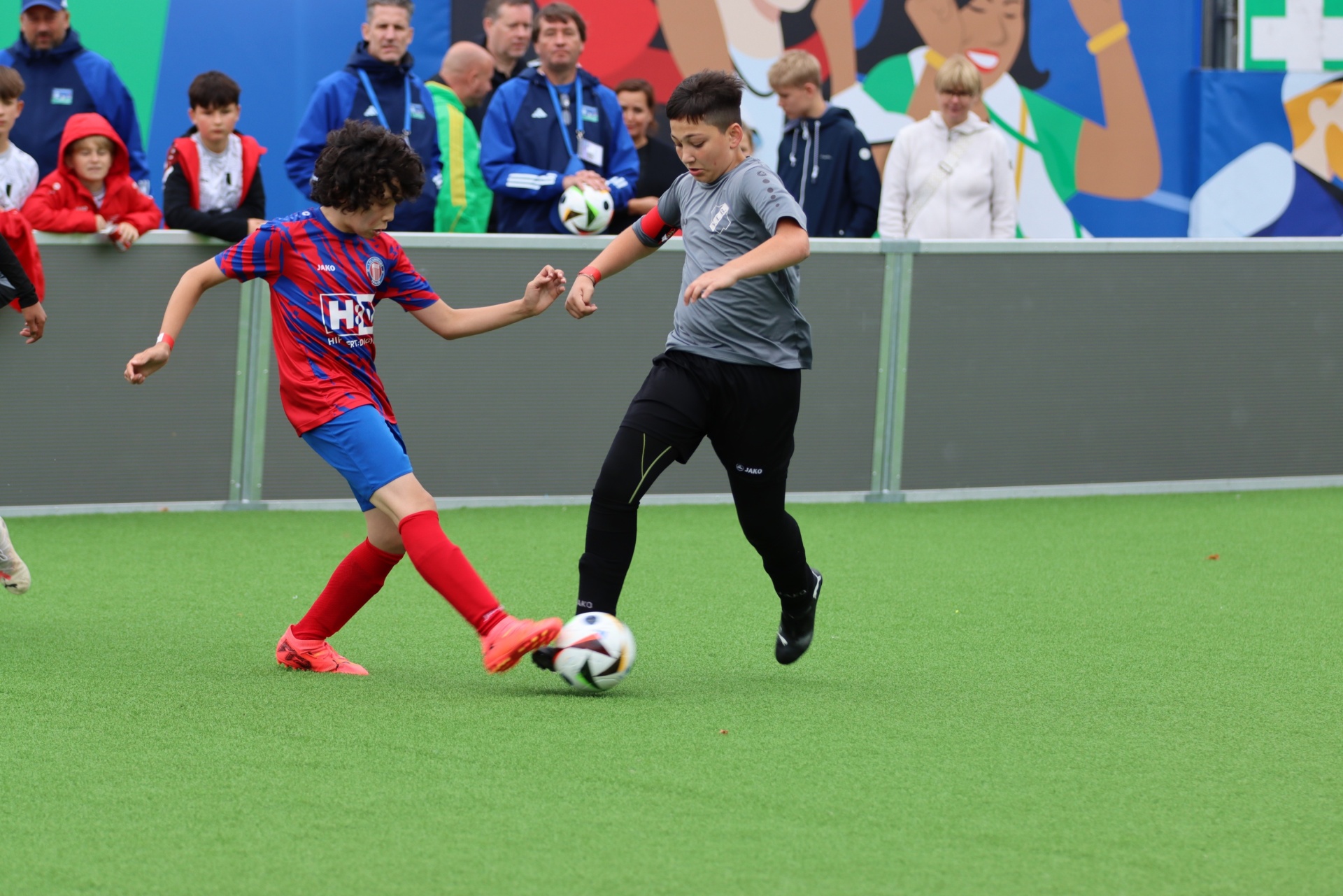 [{"clang_id":1,"value":"Zwei Jungen spielen Fußball auf einem grünen Kunstrasenplatz. Im Hintergrund sind Zuschauer und eine bunte Wand zu sehen."}]