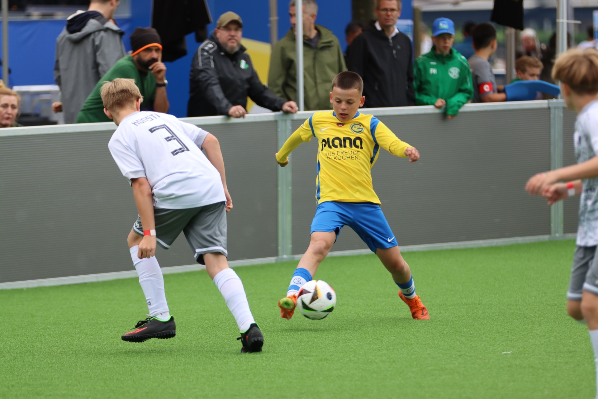 [{"clang_id":1,"value":"Zwei Fußballspieler im Spiel auf grünem Rasen. Links steht ein Spieler mit weißem Trikot, rechts dribbelt ein Spieler in blauem Trikot mit gelben Akzenten den Ball. Im Hintergrund Zuschauer."}]