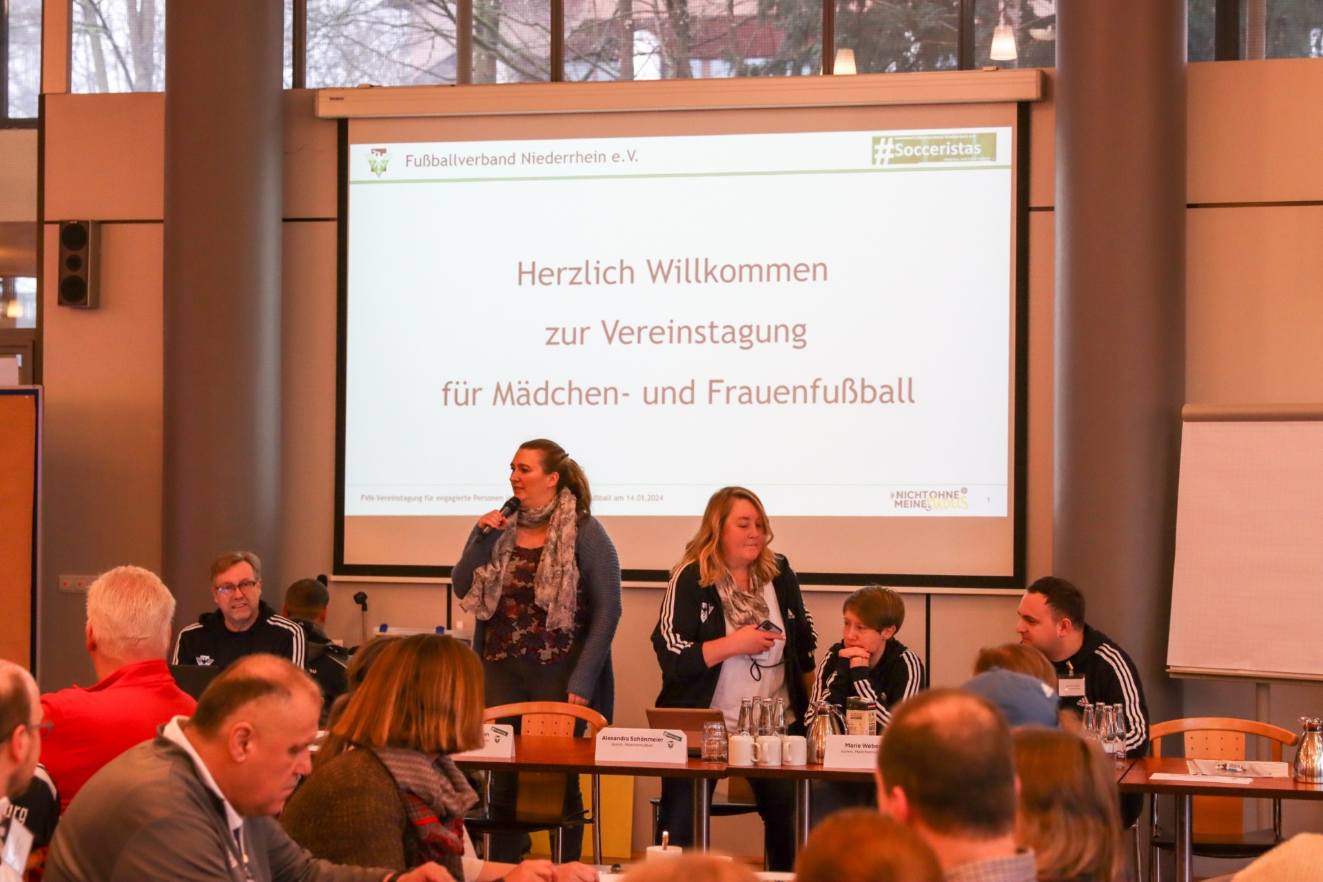 [{"clang_id":1,"value":"Versammlung in einem Raum. Zwei Frauen stehen vor einer Projektion mit Text zur Mädchen- und Frauenfußballveranstaltung. Sitzende Personen im Publikum."}]