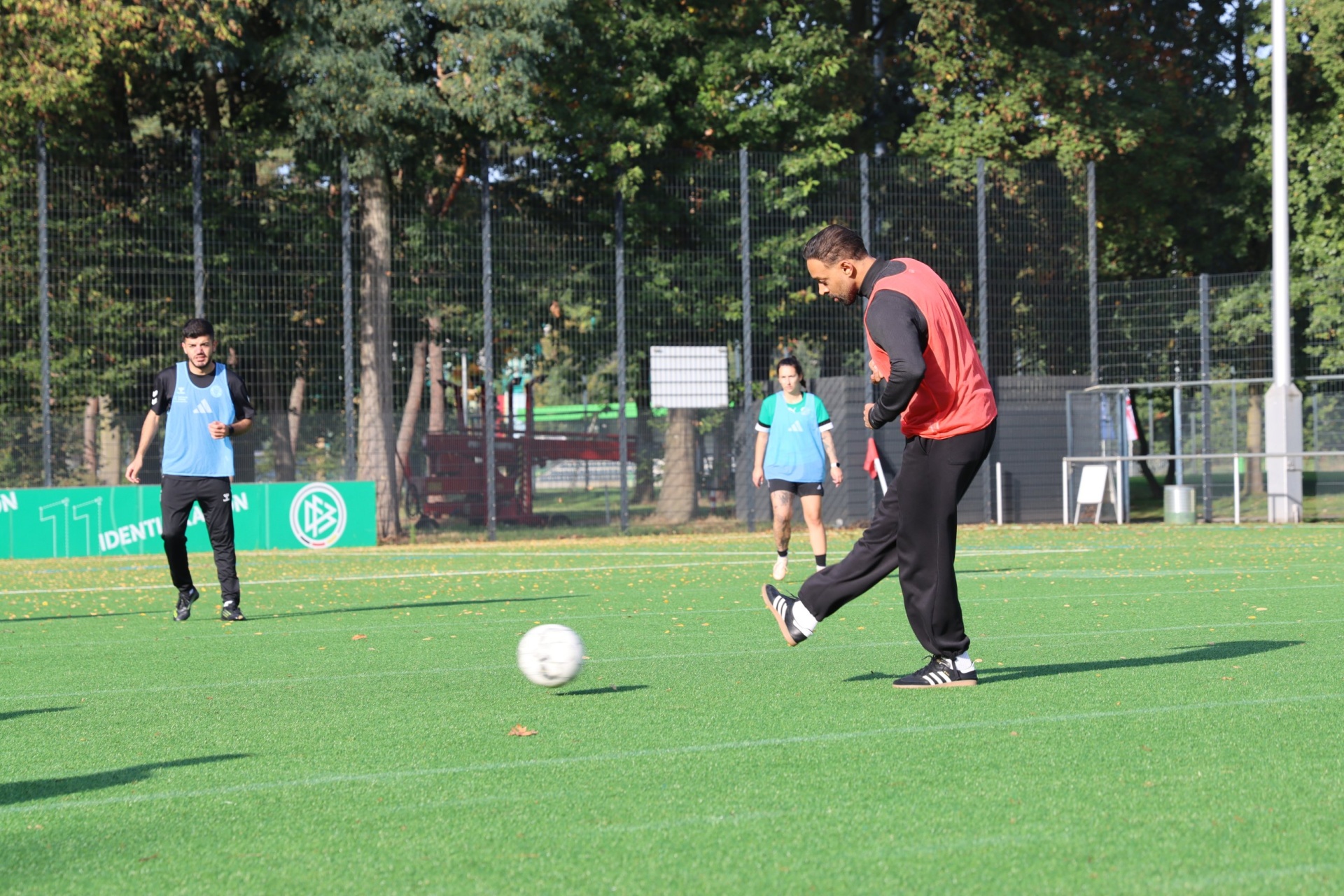 Drei Personen spielen Fußball auf einem grünen Kunstrasenplatz. Ein Mann schießt auf den Ball, zwei Jugendliche stehen abseits. Im Hintergrund ein Zaun und Bäume.