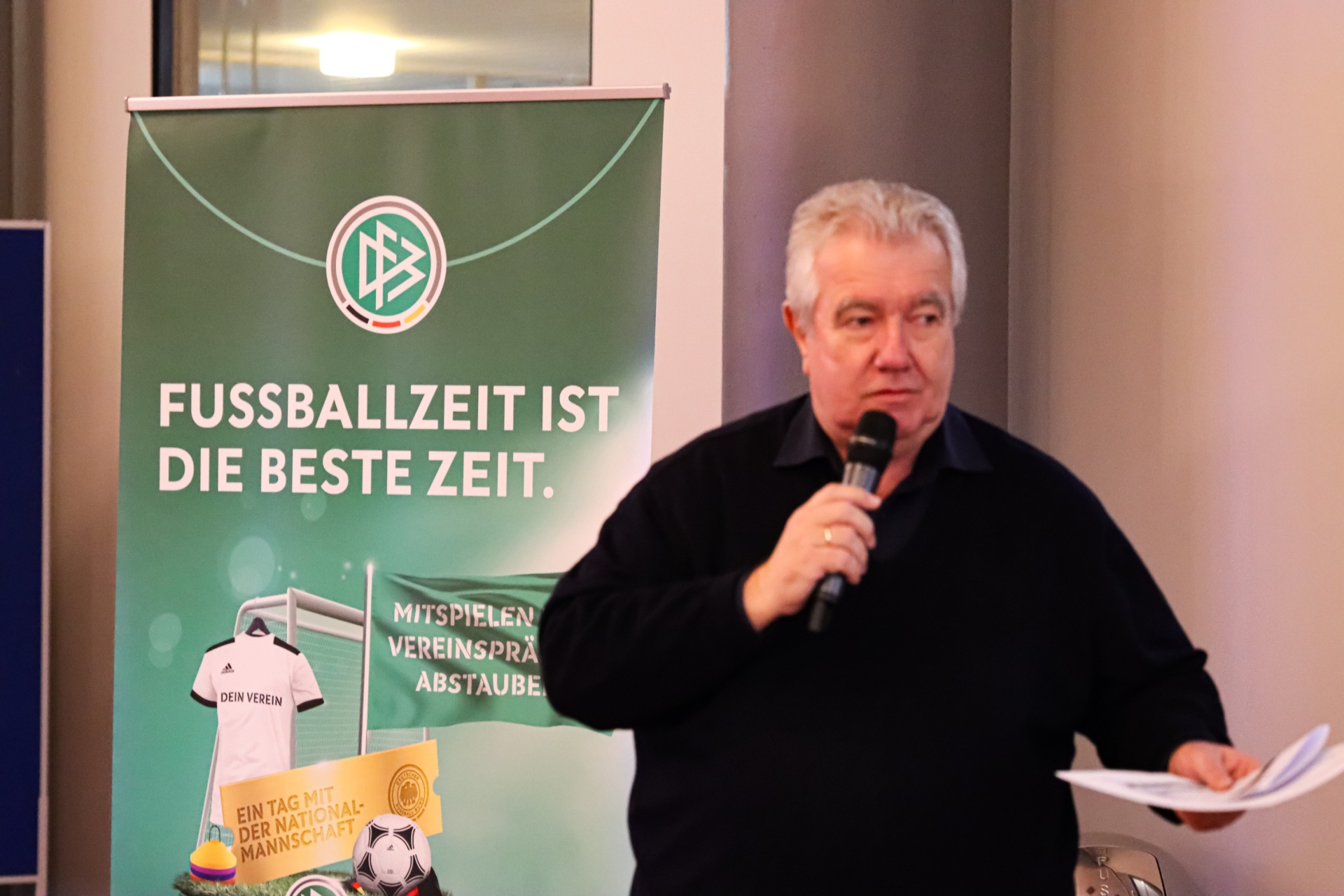[{"clang_id":1,"value":"Mann mit Mikrofon vor Banner mit Fußballmotiv und Schriftzug \"FUSSBALLZEIT IST DIE BESTE ZEIT\". Im Hintergrund Fenster und Personen."}]