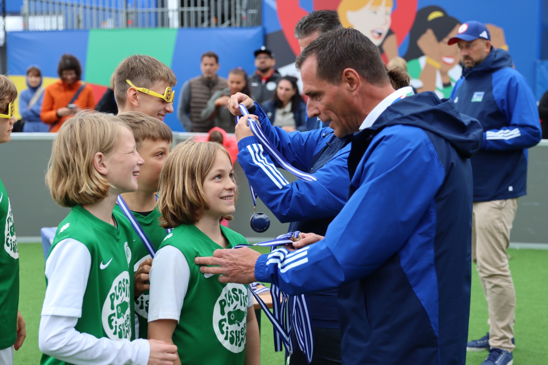 [{"clang_id":1,"value":"Männliche Person dekoriert Kinder mit Medaillen auf einem Fußballfeld. Im Hintergrund Zuschauer und bunte Wandgestaltung."}]