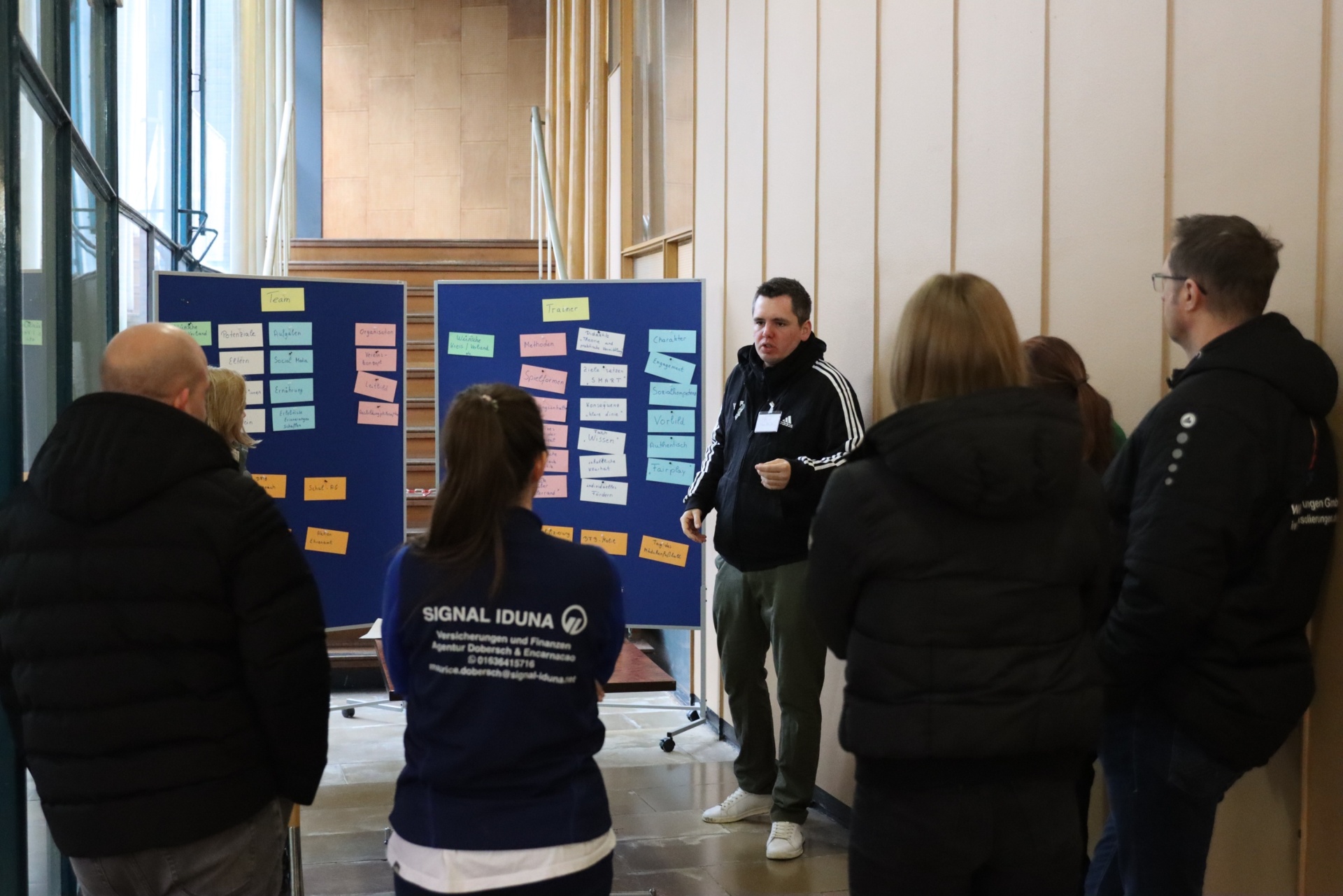 [{"clang_id":1,"value":"Gruppendiskussion vor einer Pinnwand mit Haftnotizen. Ein Mann erklärt, andere Personen hören zu. Im Hintergrund Fenster und Innenarchitektur."}]