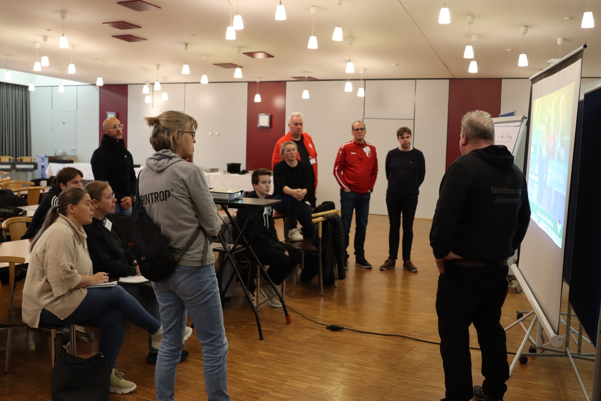 [{"clang_id":1,"value":"Raum mit etwa 15 Personen bei einer Präsentation. Ein Mann steht vor einem Projektor und spricht, andere sitzen oder stehen und schauen zu."}]