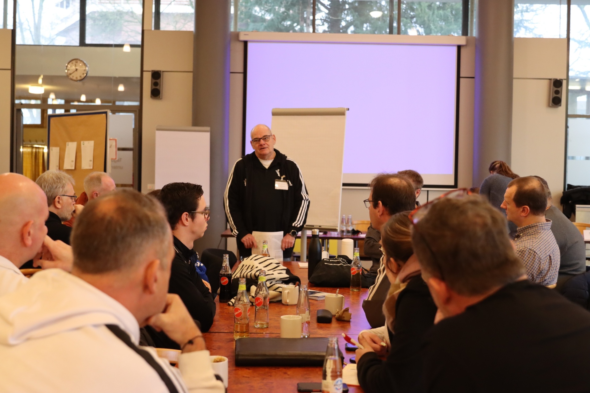 [{"clang_id":1,"value":"Konferenzraum mit Sprecher vor Publikum. Runde Tische mit Teilnehmern, Notizen und Getränken. Helle Fenster im Hintergrund."}]