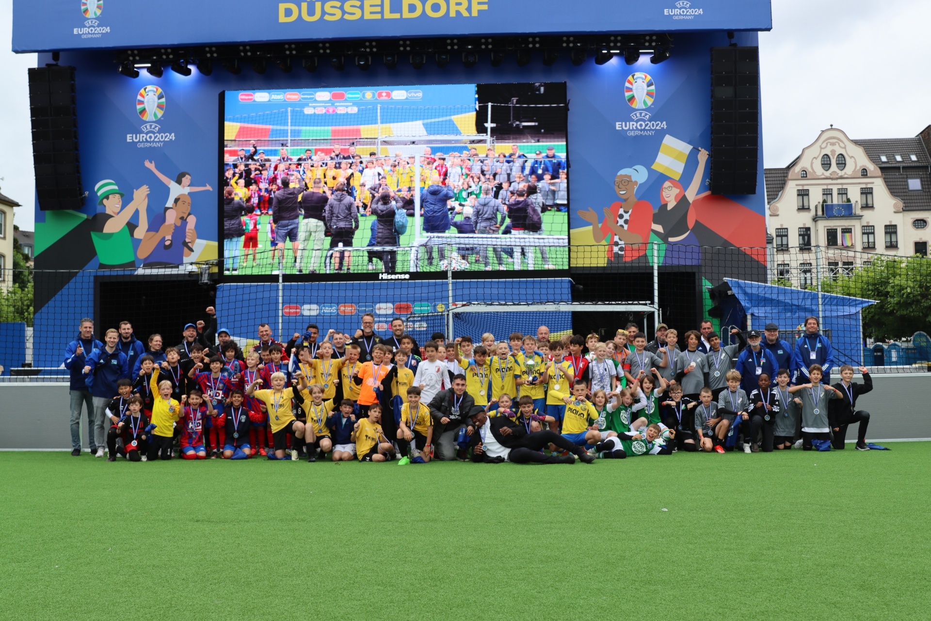 [{"clang_id":1,"value":"Fußballfeld mit vielen Kindern und Betreuern vor einer großen Leinwand mit dem Logo \"EURO 2024\" und einer Spielszene. Düsseldorf im Hintergrund."}]