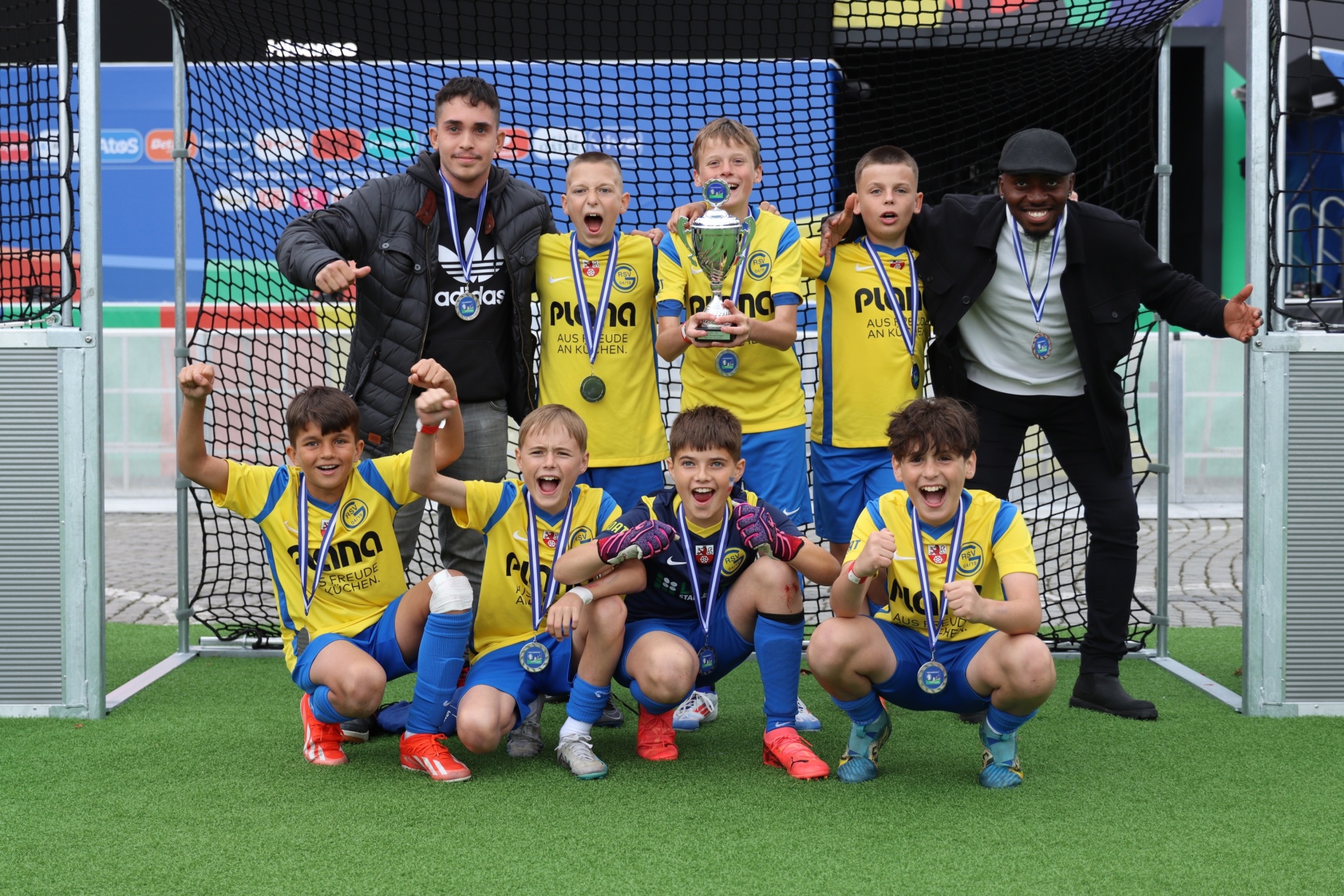 [{"clang_id":1,"value":"Fußballmannschaft mit Trainer und Betreuer posiert mit Pokal auf grünem Kunstrasen. Kinder tragen gelbe Trikots mit blauem Detail und Medaillen."}]