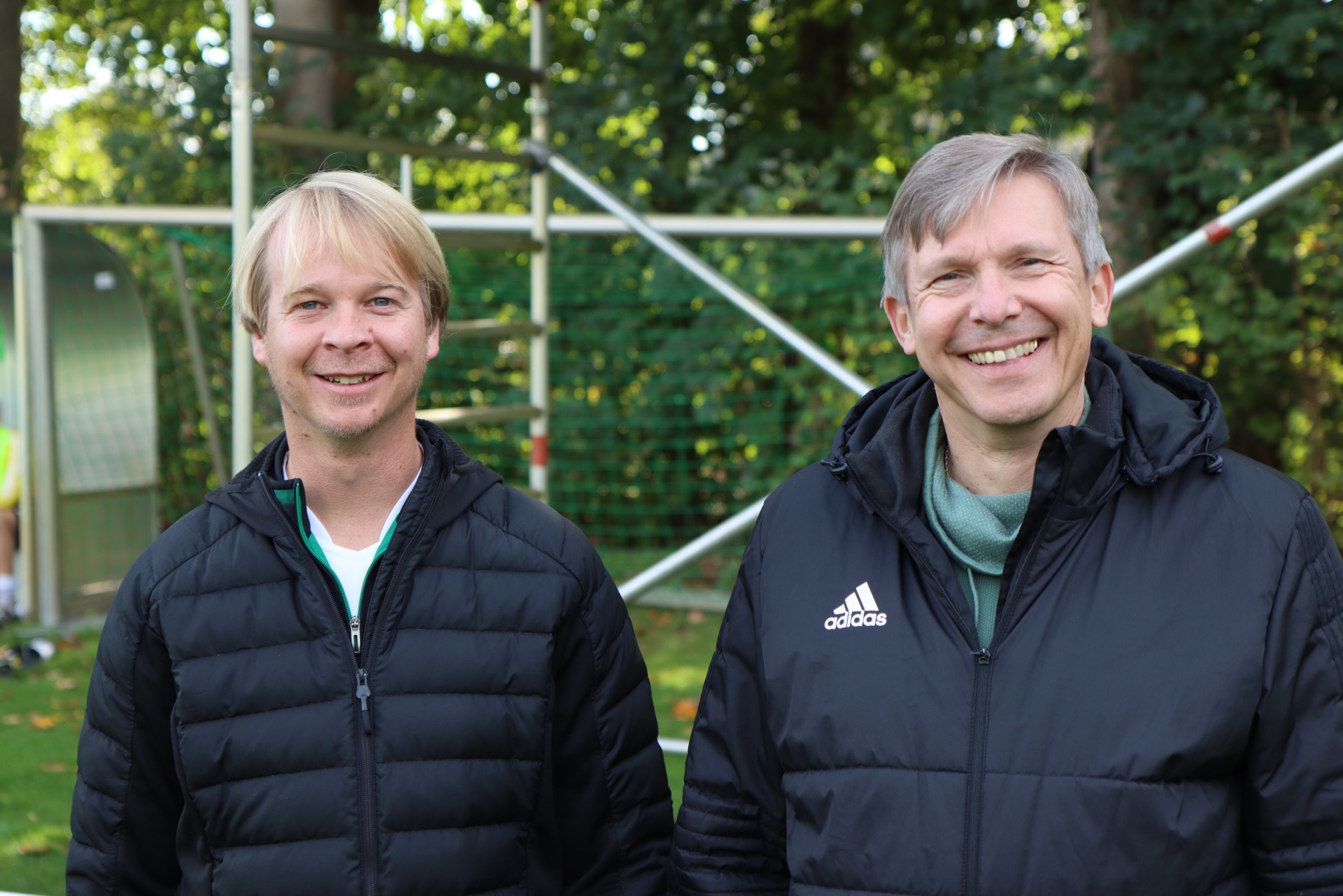 [{"clang_id":1,"value":"Zwei lächelnde Männer, beide mit blondem Haar und dunklen Jacken, stehen nebeneinander vor einem Tennisnetz und grünem Hintergrund."}]