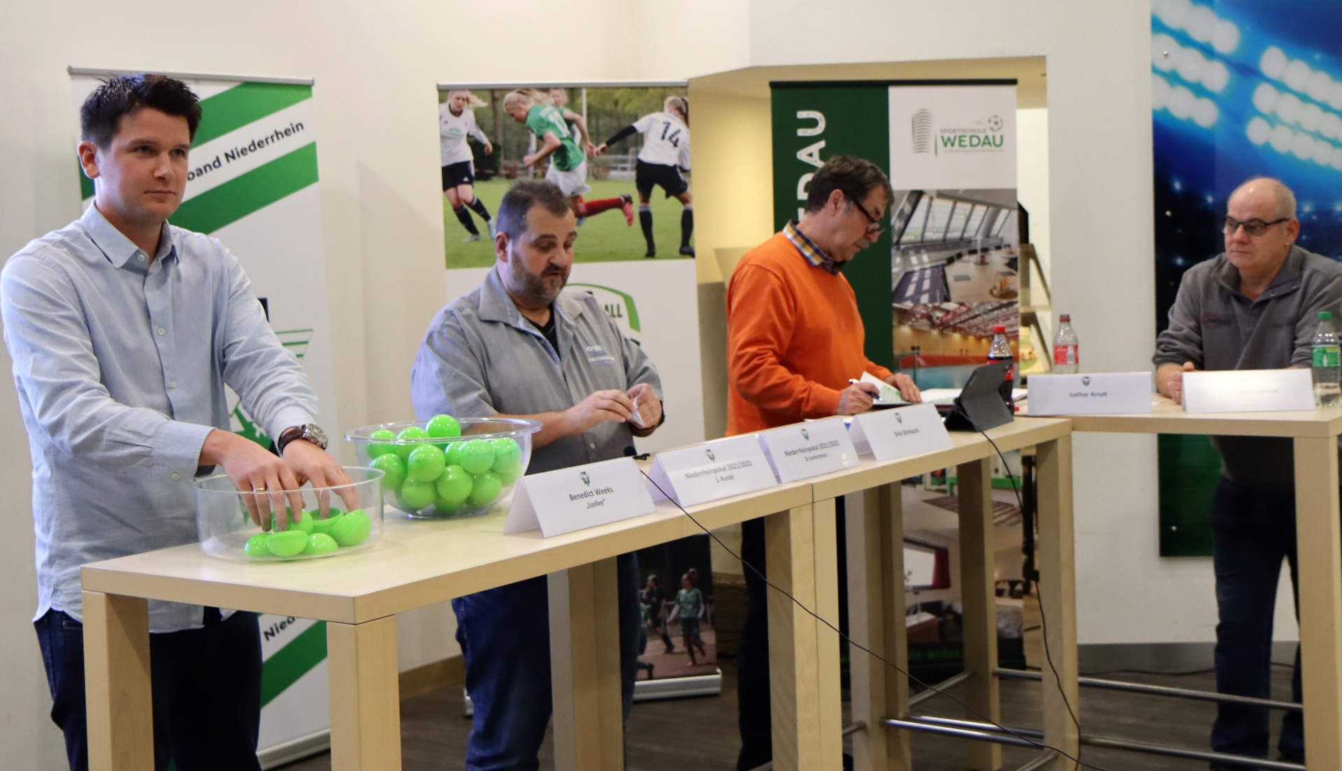 [{"clang_id":1,"value":"Fünf Männer sitzen an einem Tisch hinter dem Tennisbälle und Namensschildern stehen. Im Hintergrund eine Sportanlage und Banner."}]